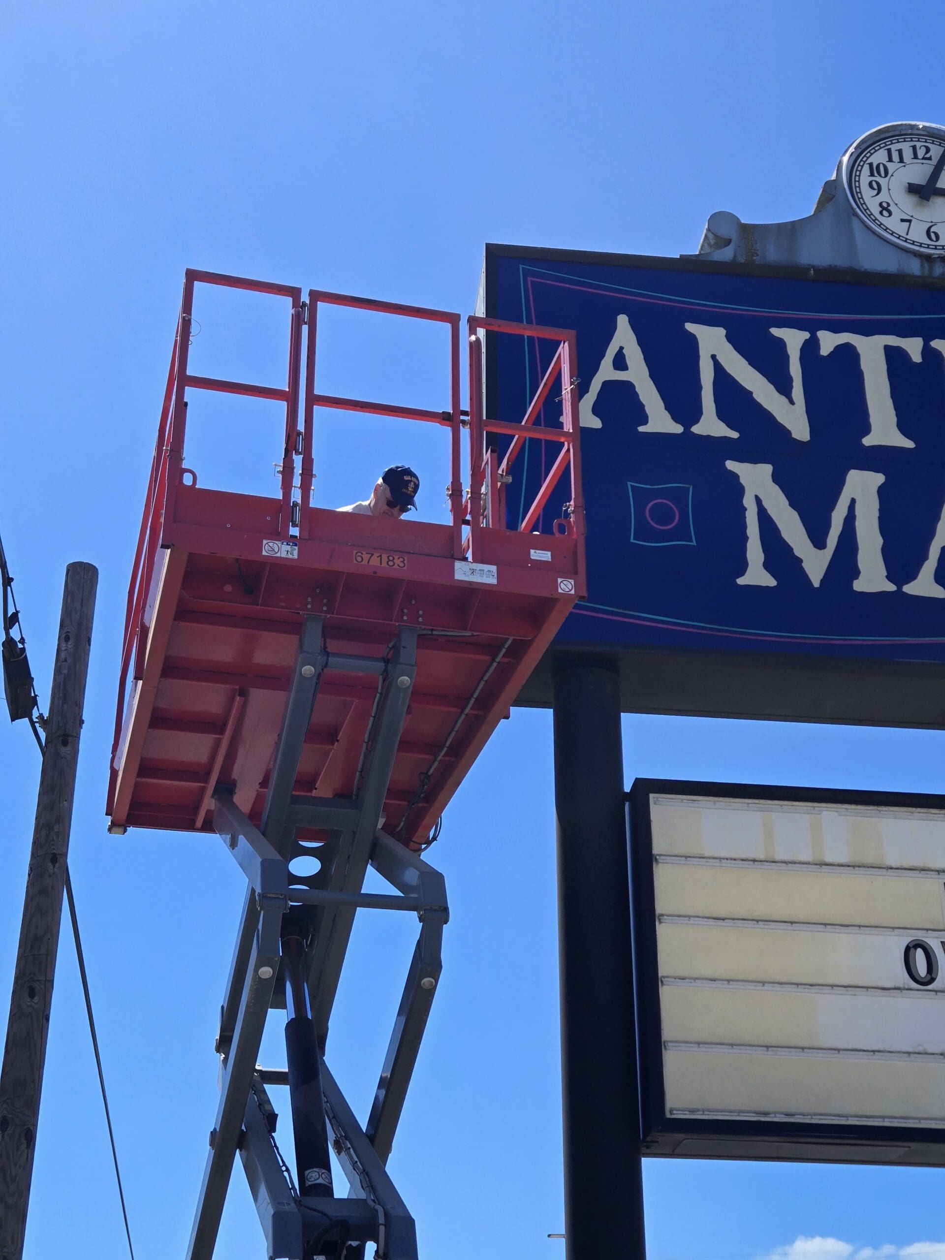 The orange SkyJack scissor lift platform fully extended beside the Pacific Antiques Mall ANTIQUE MALL panel, with a sign technician in a dark cap crouched at the railing working at the side of the sign at Pacific Antiques Mall in Tacoma, WA.