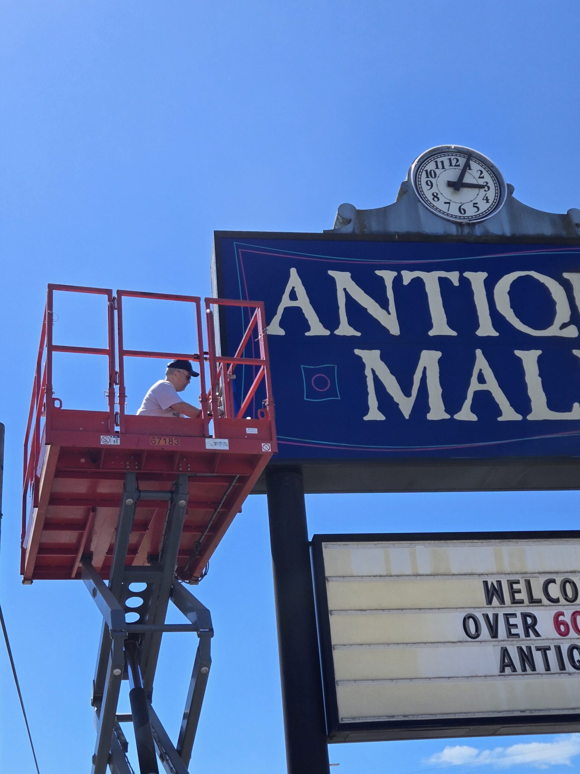 A sign technician in a white shirt and dark cap on the orange SkyJack platform working at the right edge of the deep blue Pacific Antiques Mall ANTIQUE MALL panel, with the Arabic-numeral clock above and the WELCOME OVER 60000 ANTIQUES marquee partly visible below at Pacific Antiques Mall in Tacoma, WA.