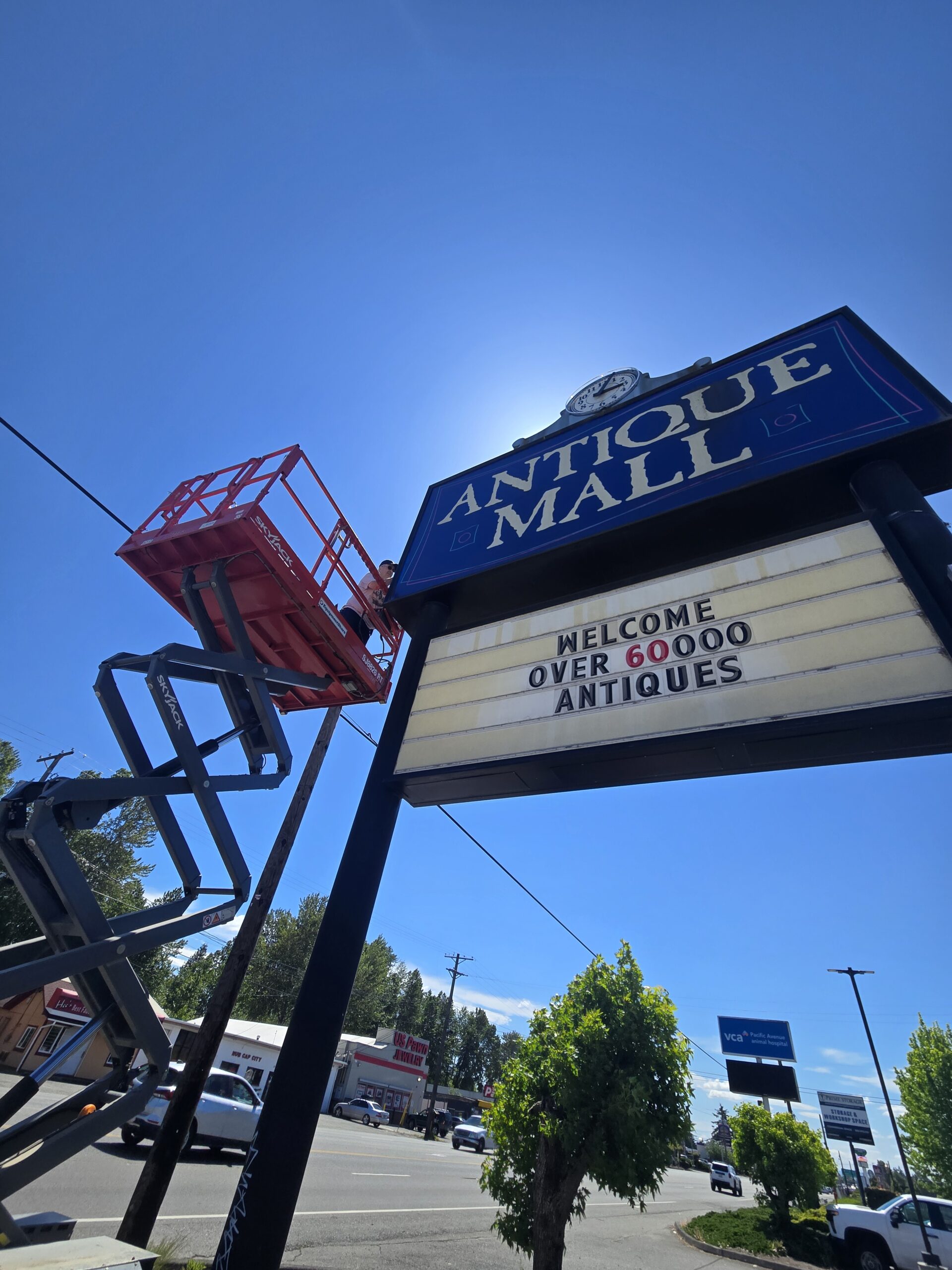 Wide sun-backed view of the Pacific Antiques Mall pylon sign on Pacific Avenue, with the ANTIQUE MALL panel and clock at top, the WELCOME OVER 60000 ANTIQUES marquee below, the SkyJack scissor lift extended on the left with the technician on the platform, and Pacific Avenue businesses including the VCA Pacific Avenue Animal Hospital visible in the background at Pacific Antiques Mall in Tacoma, WA.