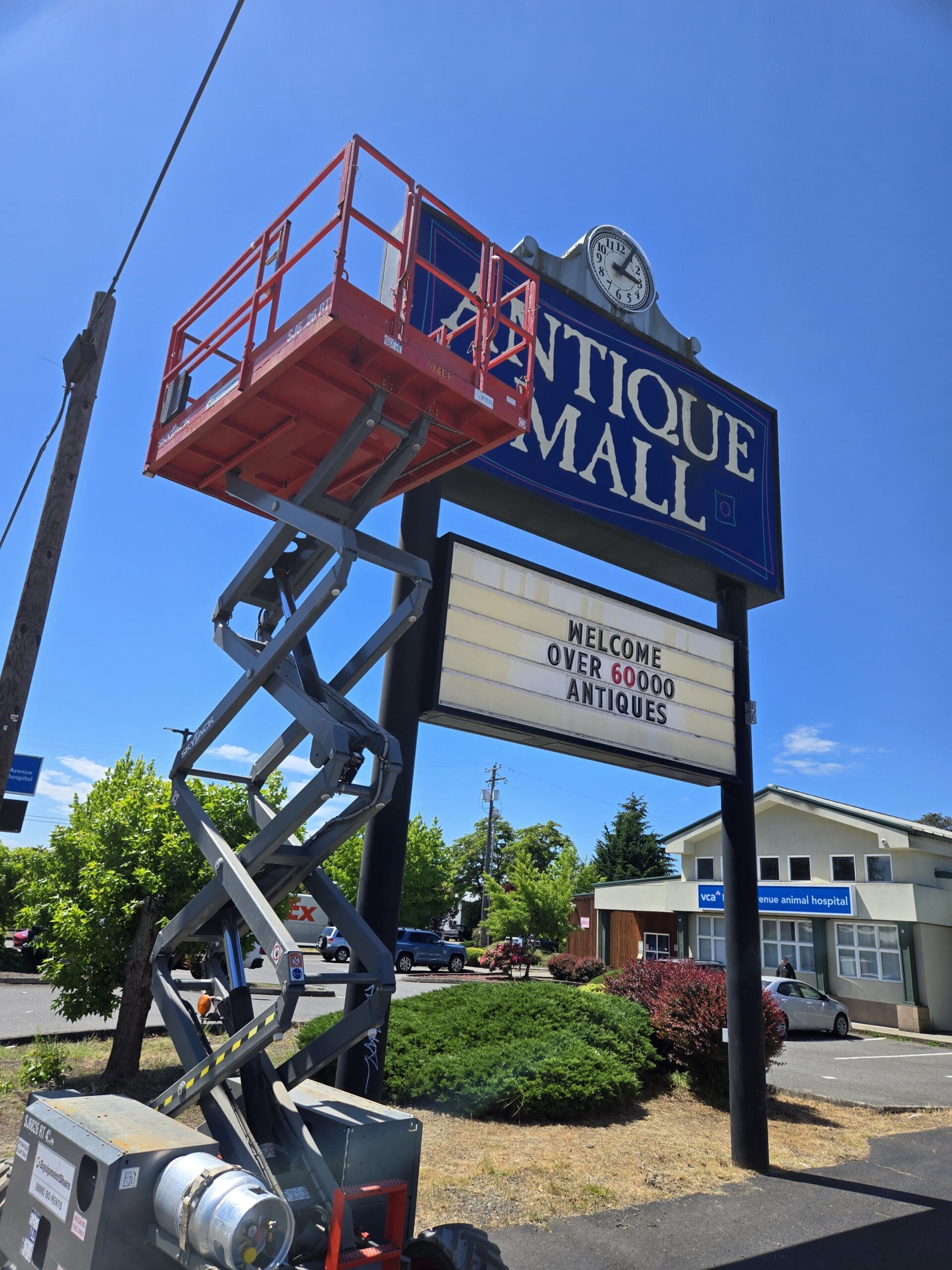 Side view of the Pacific Antiques Mall pylon sign with the deep blue ANTIQUE MALL panel and clock at top and the WELCOME OVER 60000 ANTIQUES marquee below, beside the SkyJack scissor lift fully extended with the platform empty, and the VCA Pacific Avenue Animal Hospital and Pacific Avenue parking area in the background at Pacific Antiques Mall in Tacoma, WA.
