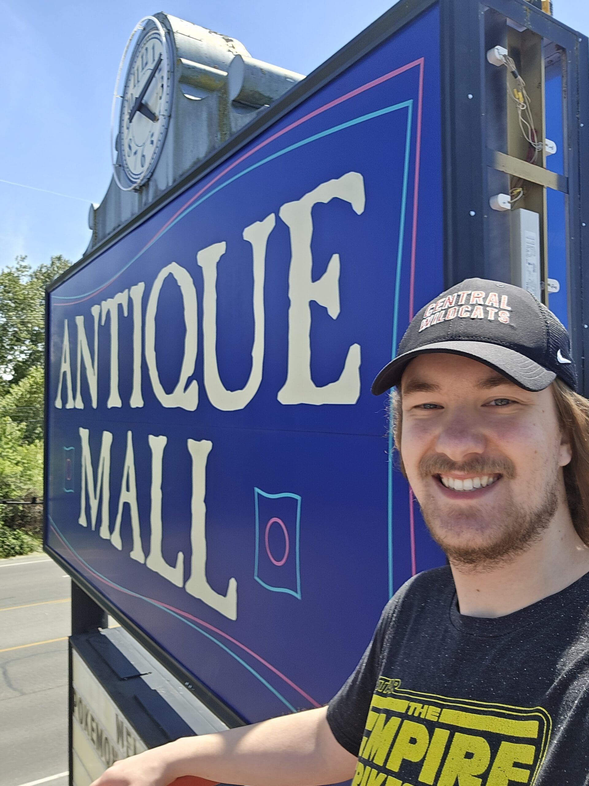 Close daytime view of the Pacific Antiques Mall ANTIQUE MALL sign panel from the lift platform, with the painted white and pink-coral lettering on deep blue and the side electrical access panel open to show the white sockets and red and yellow internal wiring, with a Pacific Antiques Mall team member smiling beside the sign in a Central Wildcats cap and an Empire Strikes Back T-shirt at Pacific Antiques Mall in Tacoma, WA.