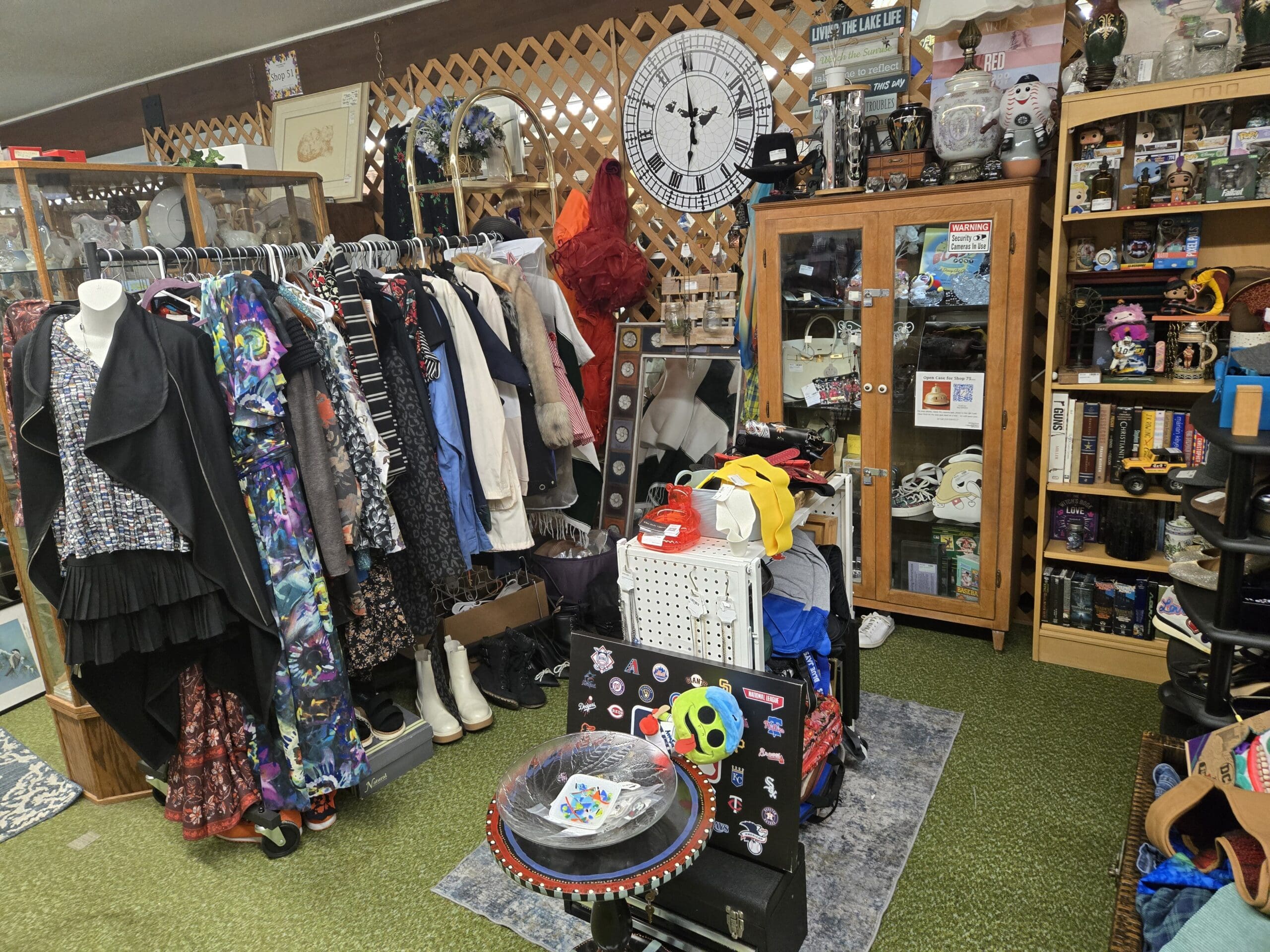 Wide view of Shop 75 with a packed clothing rack of vintage dresses and patterned blouses on the left, a headless mannequin in a black draped cardigan over a black-and-white check sleeveless top with a pleated skirt, a large Big Ben style wall clock with a Peter Pan flying scene on the lattice wall above, glass-fronted wood cabinets of handbags and jewelry on the right, a Funko Pop and vintage decor shelf at the far right, and a round red folk-art painted side table with a glass leaf dish and an MLB National League logo board in the center foreground at Shop 75, Pacific Antiques Mall in Tacoma, WA.