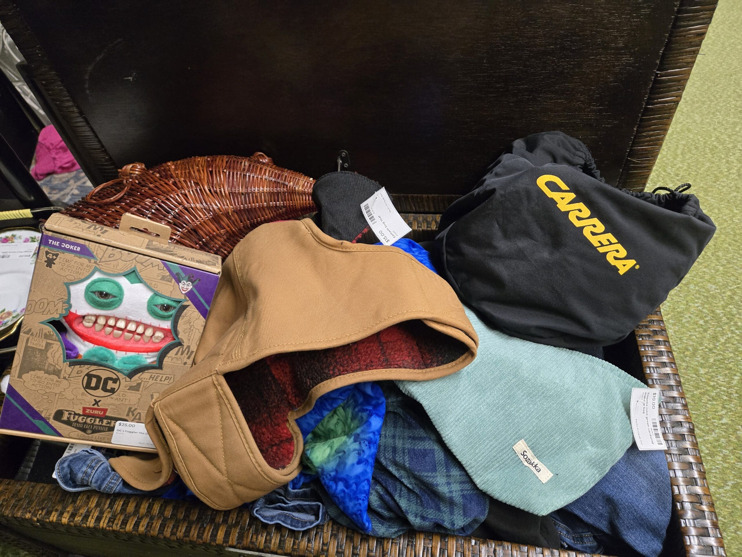 Open wicker storage chest holding a boxed DC x Zuru Fuggler The Joker plush in original packaging, a small wicker basket, a tan Carhartt style cropped wraparound hood, a black drawstring CARRERA sunglasses pouch, a red-and-black plaid fleece scarf, a teal soft pouch labeled Sazika, and folded denim at Shop 75, Pacific Antiques Mall in Tacoma, WA.