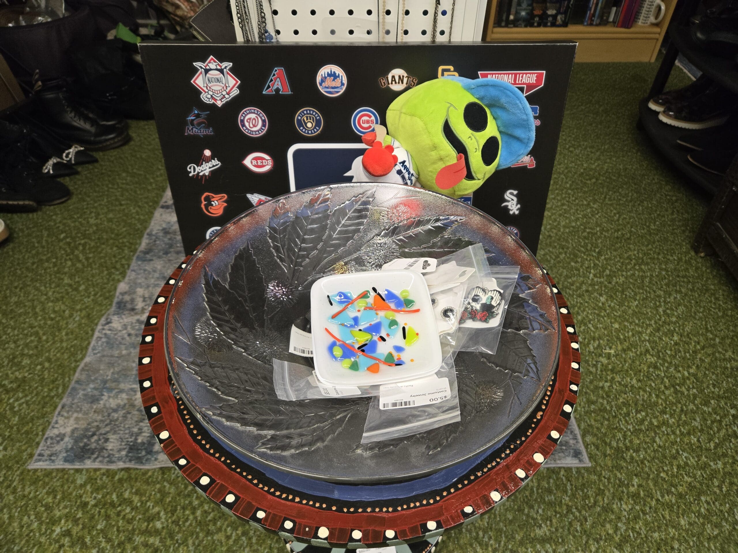 Round folk-art painted side table with red and white dotted trim holding a glass leaf-shaped serving dish, a small white ceramic tray of brightly colored fused-glass earrings, and a bagged costume jewelry set, with a National League MLB logo board behind featuring Phillies, Diamondbacks, Mets, Giants, Marlins, Brewers, Cubs, Dodgers, Reds, Orioles, and White Sox team marks, plus a green plush alien character on top at Shop 75, Pacific Antiques Mall in Tacoma, WA.