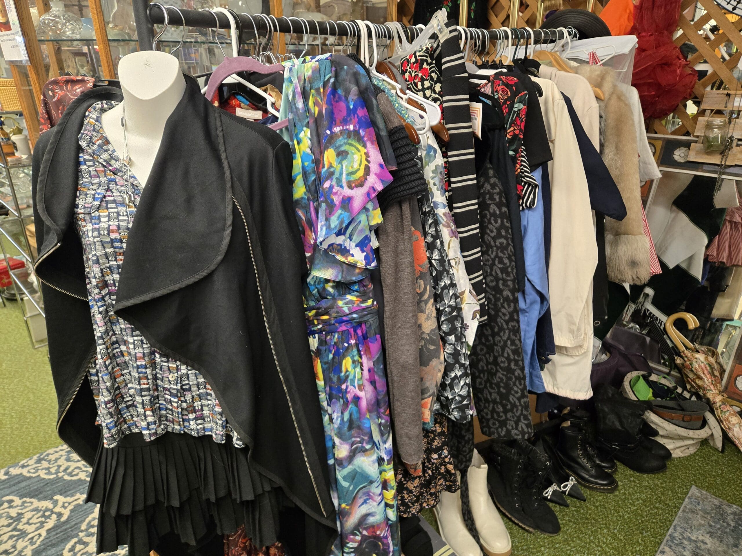 Closer view of the Shop 75 clothing rack with a white mannequin in a black draped cardigan over a herringbone sleeveless top and pleated black skirt in front, dresses and blouses behind in purple and yellow tie-dye, gray, navy, blue, black with rose floral, gray leopard, white, and a fur-collared coat, with black lace-up ankle boots, white pointed Chelsea boots, and a floral-print umbrella with curved cane handle on the floor at Shop 75, Pacific Antiques Mall in Tacoma, WA.