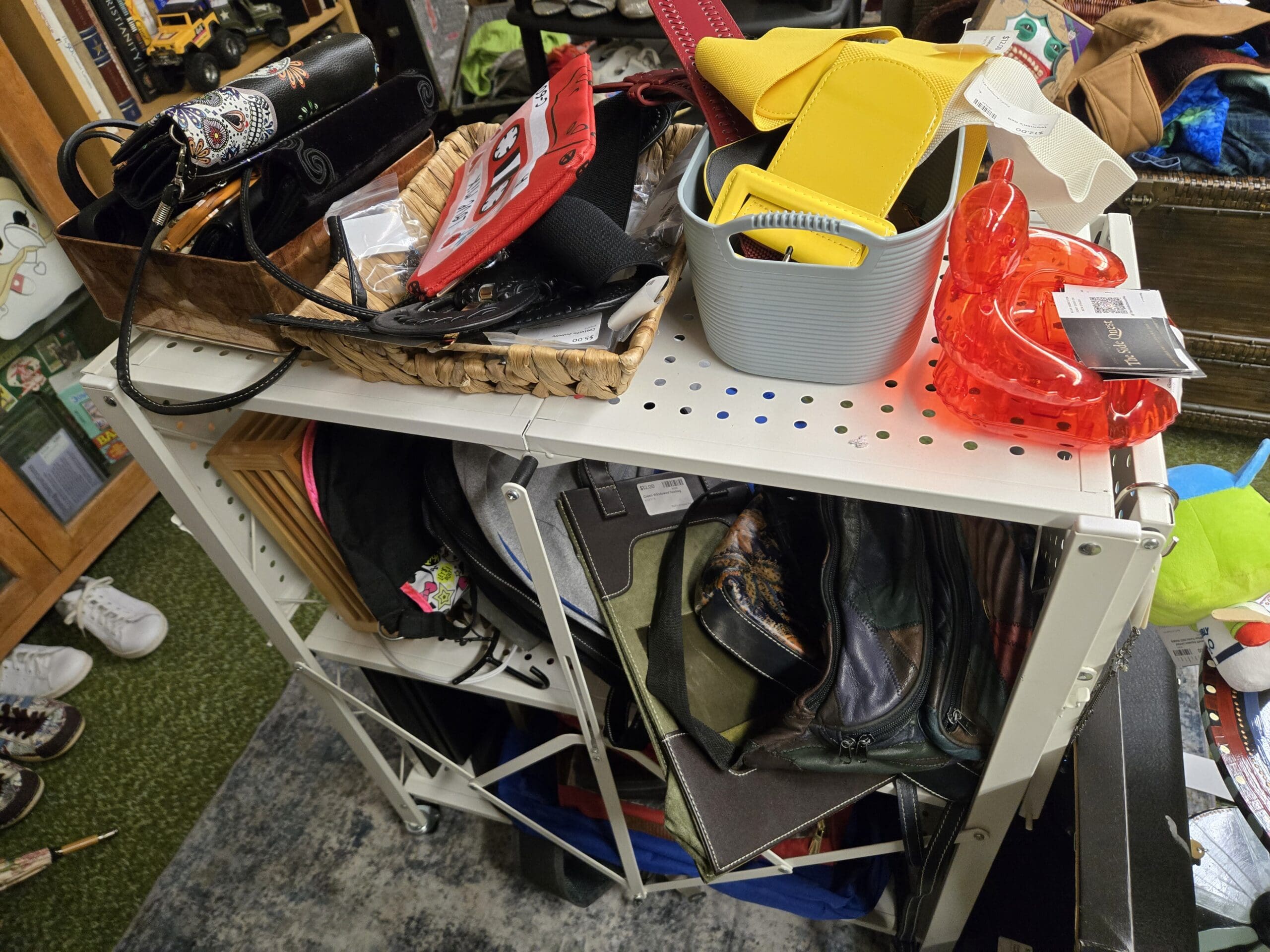 White wire shelving unit with a wicker basket on top holding a black sugar skull decorated tin, a small fan-shaped wicker piece, a yellow handbag, and bagged accessories, with a clear orange-red plastic sandal nearby, and a lower shelf packed with knee-high black leather boots, stacked handbags, a backpack, and animal-print scarves at Shop 75, Pacific Antiques Mall in Tacoma, WA.