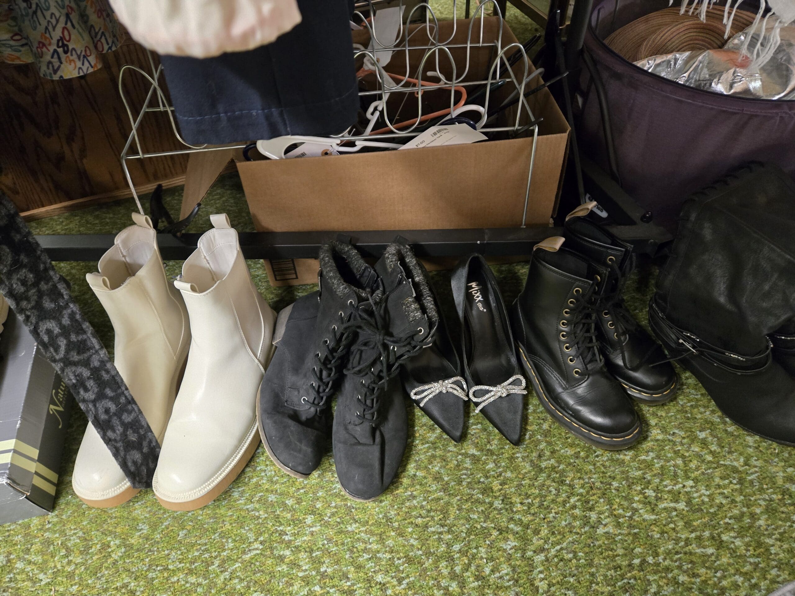 Footwear lined up on green carpet including cream pull-on Chelsea ankle boots, black suede lace-up combat boots, black pointed-toe stilettos with silver rhinestone bow toes from Mixx, and tall black leather lace-up 1460-style combat boots, in front of a cardboard box of wire hangers at Shop 75, Pacific Antiques Mall in Tacoma, WA.