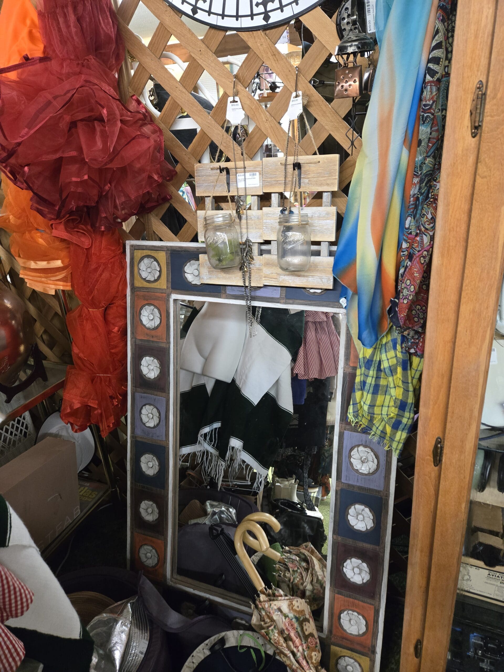Lattice corner with a decorative wood-framed standing mirror bordered by alternating blue, orange, and brown tile inserts with white pressed floral medallions, a whitewashed barnwood plaque mounted with two Ball mason jars used as a jewelry organizer, sheer red and orange ruffled wrap scarves draped on the lattice, a yellow plaid fringe scarf, and a black metal hanging lantern at Shop 75, Pacific Antiques Mall in Tacoma, WA.