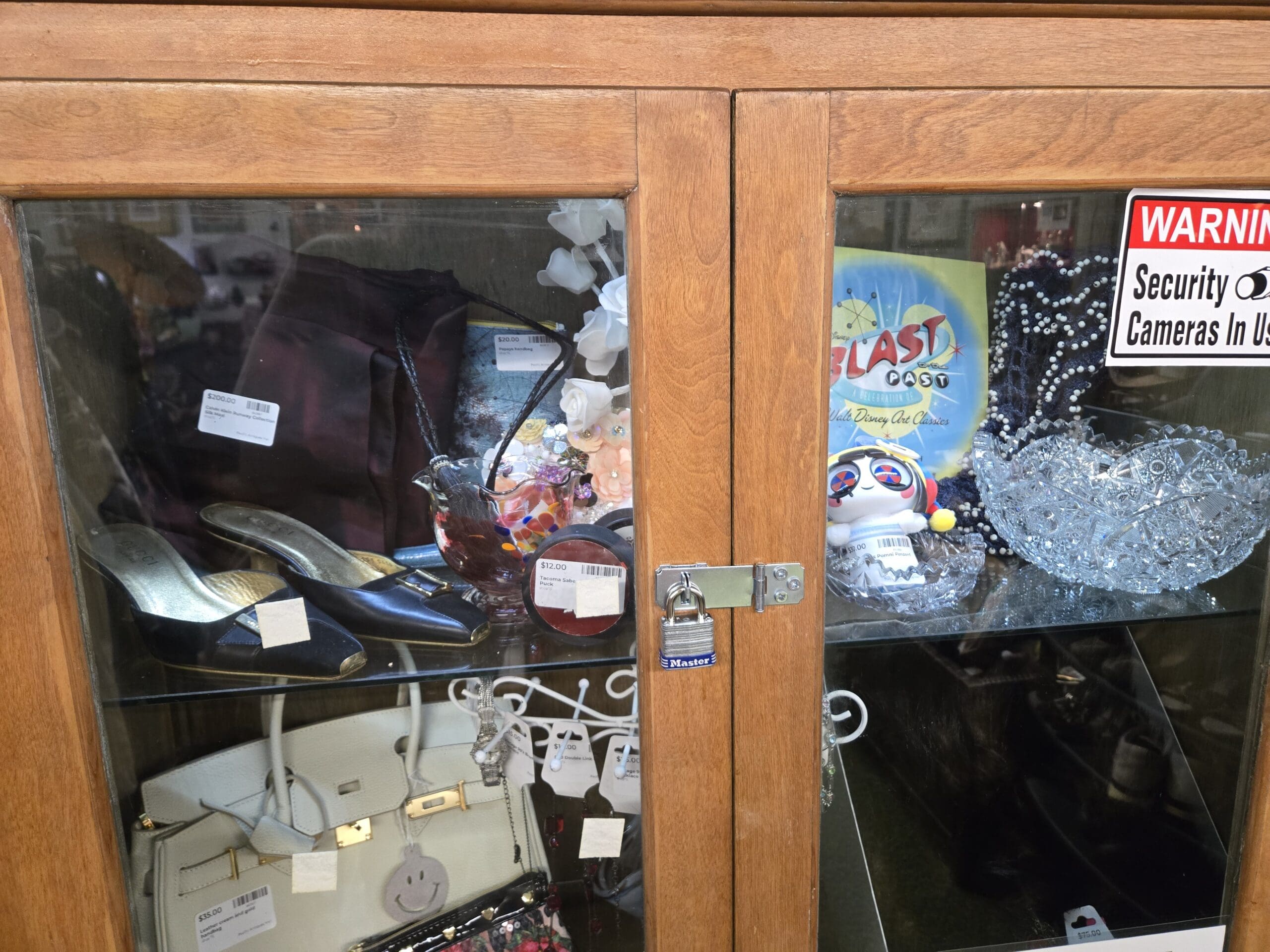 Glass-fronted wood display cabinet locked with a Master padlock, with a vintage black satin clutch, vintage pumps with white toe and gold heel detail, a plum velvet jacket, silk flower decoration, and an amber glass vase on the upper shelf, a cream leather satchel-style handbag with gold turn-lock hardware, a smiley-face charm, and sequined clutches on the lower shelf, with a Walt Disney Art Classics Blast from the Past Mickey-style figurine, a beaded crystal bib necklace, a cut crystal bowl, and a WARNING Security Cameras In Use sign on the adjacent cabinet at Shop 75, Pacific Antiques Mall in Tacoma, WA.