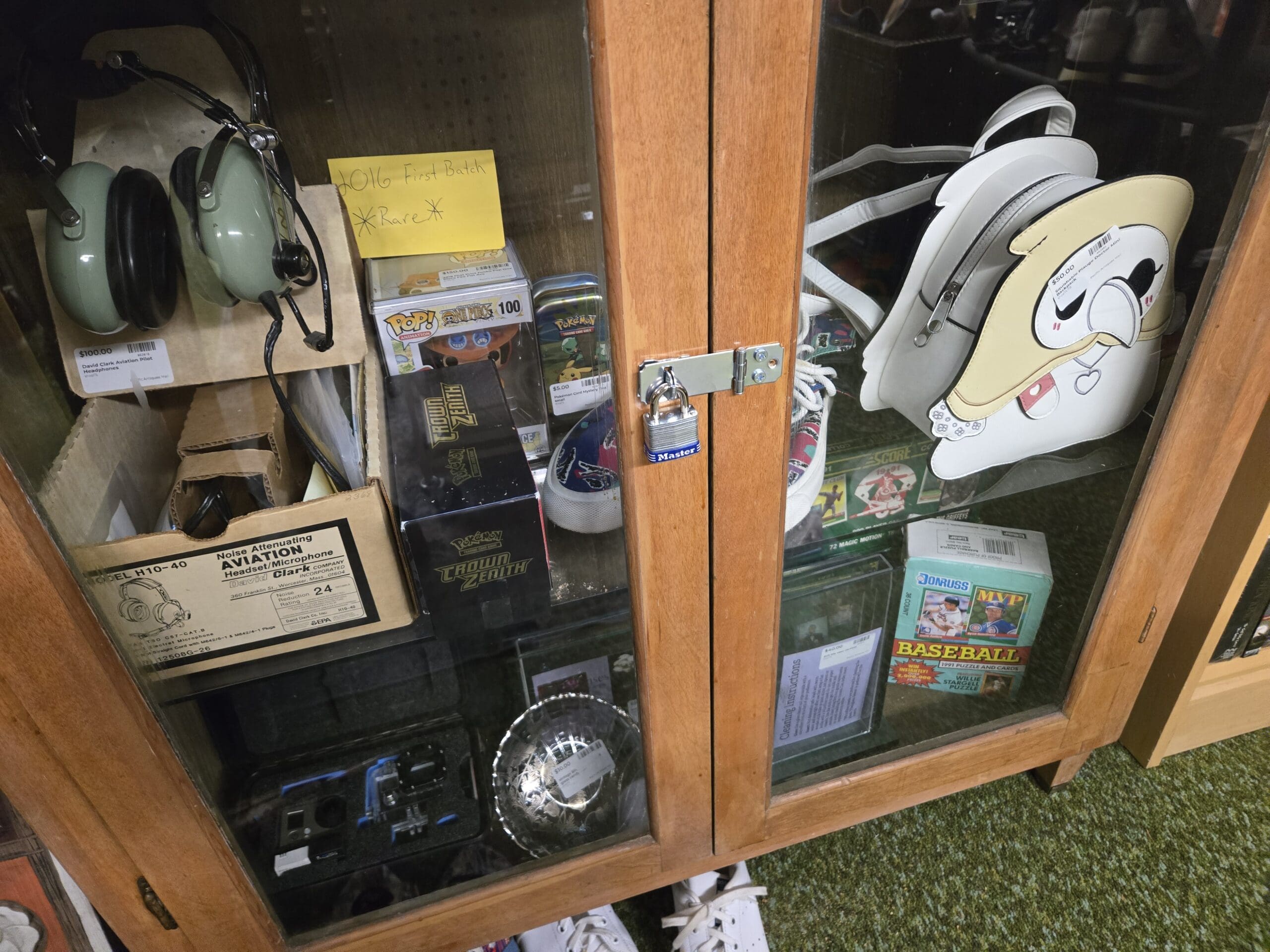 Glass-fronted wood cabinet locked with a Master padlock, with a David Clark H10-40 noise-attenuating aviation headset in sage green in its original box on the upper left, a Funko Pop Pokemon Snivy figure labeled First Batch Rare, a Pokemon Crown Zenith Elite Trainer Box, and a Pokemon tin in the center, a white novelty character mini backpack with chubby cheek face design on the right, and a 1991 Donruss MVP Baseball card box and a Score baseball card box on the lower shelf at Shop 75, Pacific Antiques Mall in Tacoma, WA.