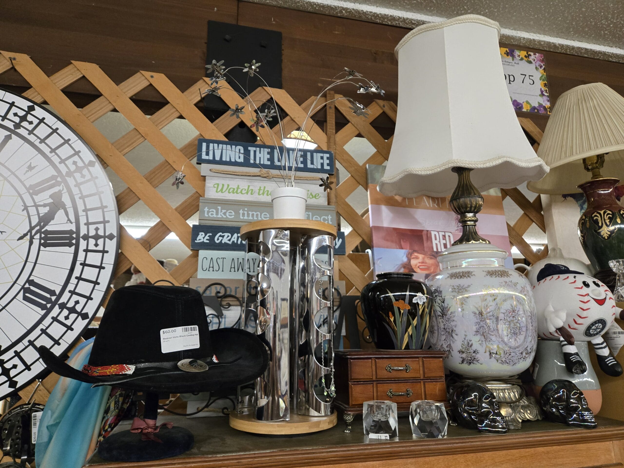 Display shelf with a partial view of the Big Ben Peter Pan wall clock on the left, a black western hat with a beaded band, a chrome cylindrical sunglasses display tower, a stacked Living the Lake Life wood-block inspirational sign reading watch the sunrise, take time to reflect, be grateful, and cast away, a black Japanese-style vase with painted gold florals, a cream and gold floral lidded ginger jar, two crystal cut paperweights, two ceramic sugar skull figurines in silver and black, a vintage table lamp with a cream pleated shade, and a Mariners baseball plush character at Shop 75, Pacific Antiques Mall in Tacoma, WA.