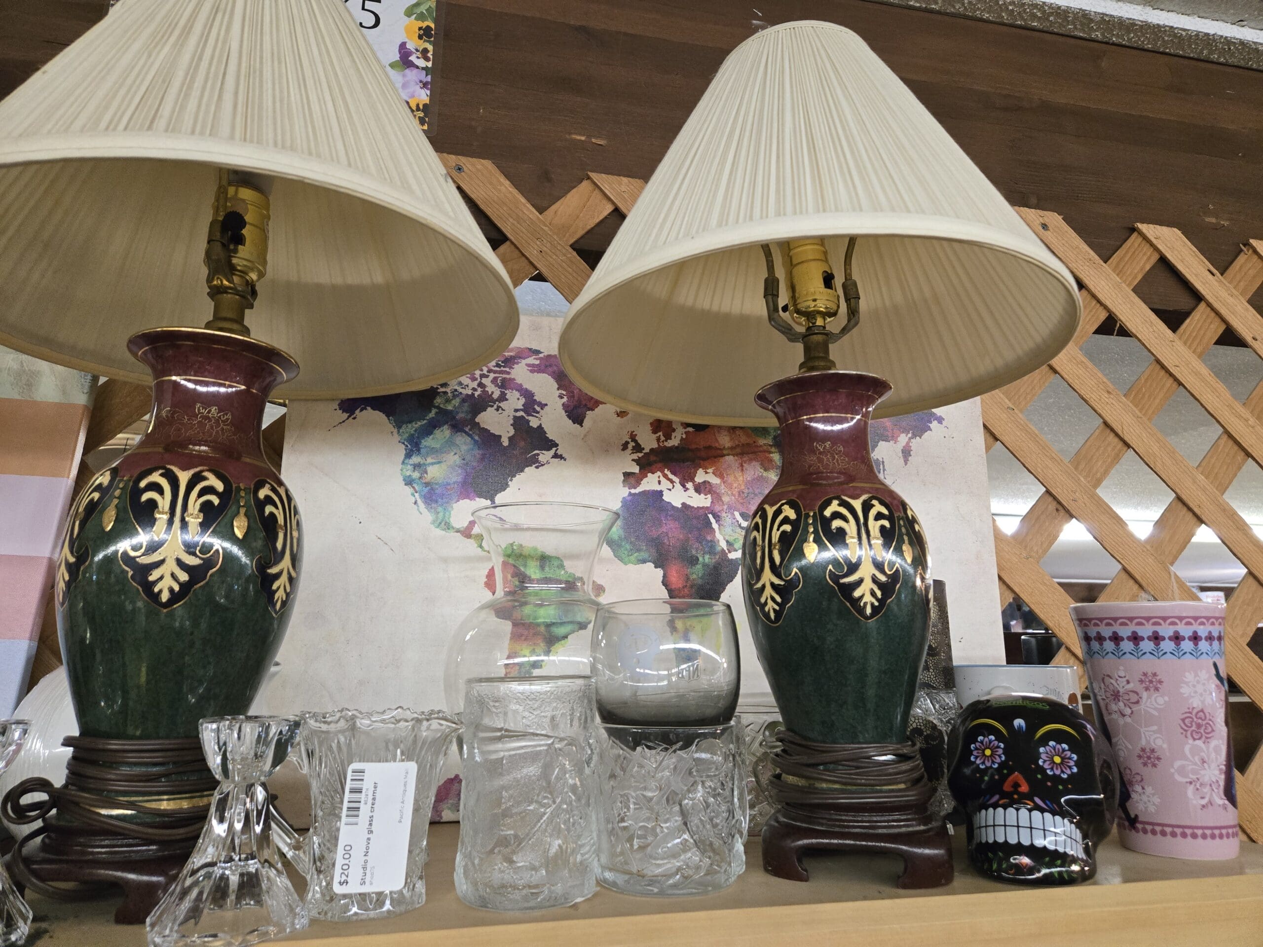Shelf with a pair of matching emerald green and burgundy table lamps decorated with gold fleur-de-lis scrollwork on small wood pedestals with cream pleated shades, three clear textured glass tumblers including a Studio Nova glass creamer, a clear hurricane vase, a black painted ceramic sugar skull with white floral detail, and a pink-and-white floral tumbler, set against a watercolor world map artwork backdrop at Shop 75, Pacific Antiques Mall in Tacoma, WA.