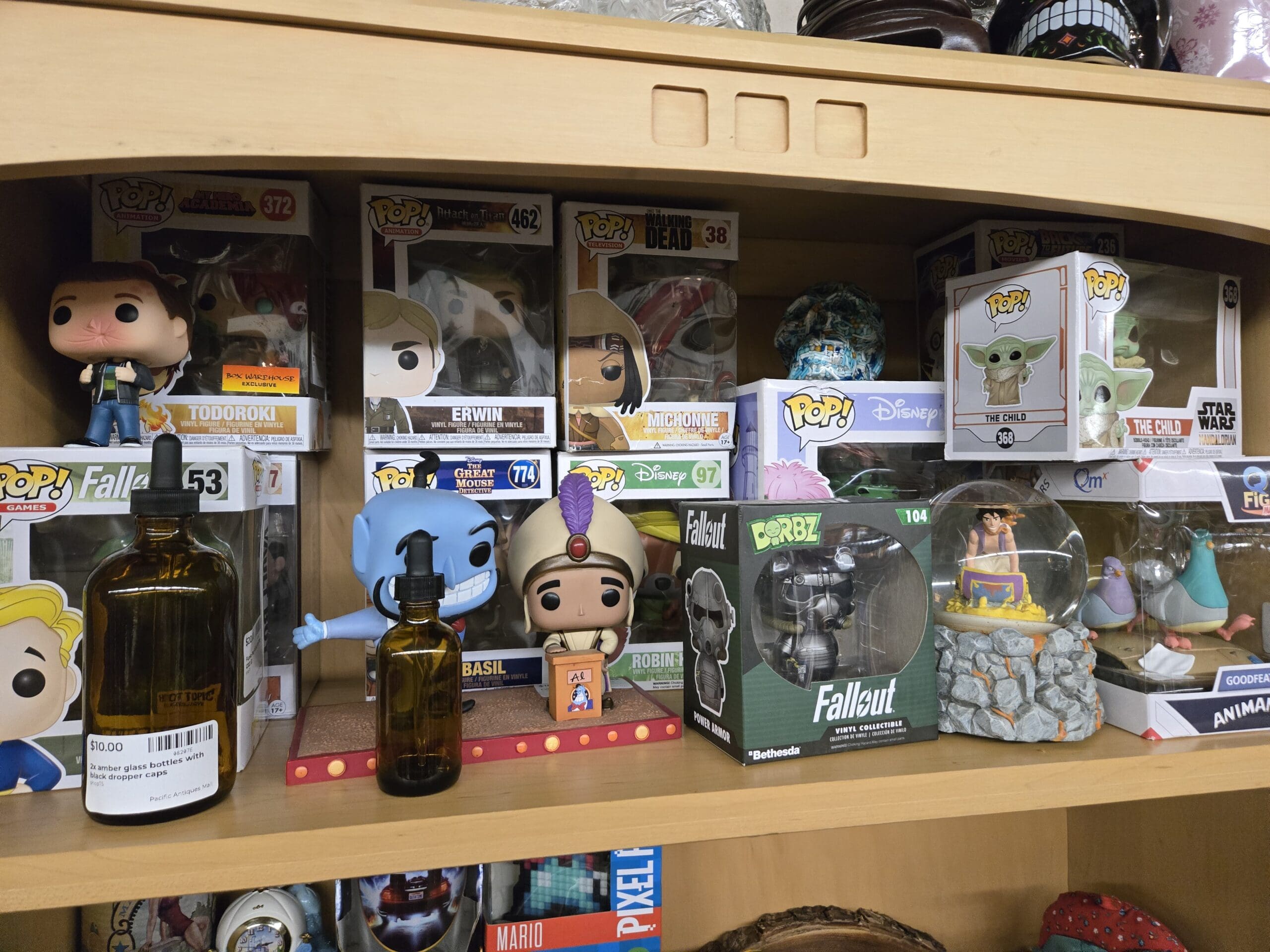 Funko Pop figure shelf with boxed Pop Animation Todoroki #372 from My Hero Academia, Pop Animation Erwin #462 from Attack on Titan, Pop Television Walking Dead Michonne #38, and Pop Star Wars The Child #368 from The Mandalorian on the upper row, with an out-of-box Genie and Prince Ali from Aladdin, a Disney Great Mouse Detective Basil #774, a Disney Robin Hood Robin #97, a Pop Games Fallout #53 Vault Boy, a Fallout Dorbz #104 Power Armor, an Aladdin snow globe, and two amber glass dropper bottles in front at Shop 75, Pacific Antiques Mall in Tacoma, WA.