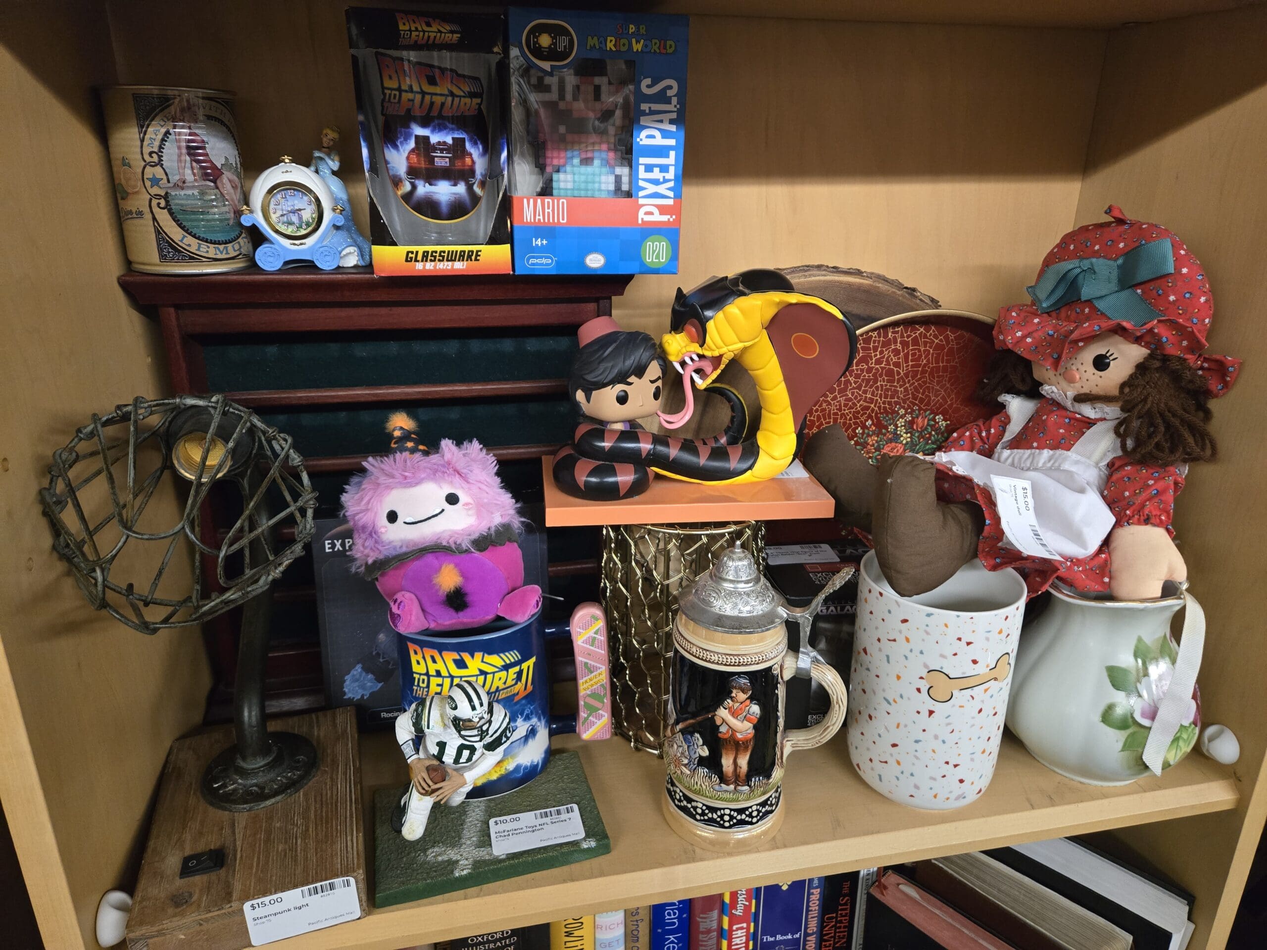 Eclectic shelf with a vintage wire cage steampunk-style lamp, a Cinderella style blue carriage clock, a boxed Back to the Future glassware pint glass, a boxed Pixel Pals Super Mario World #020 light-up figure, a pink fuzzy Fuggler plush on a Back to the Future Part II themed cup, a Funko style Pop figure of a person with a yellow cobra, a vintage German beer stein with painted fly-fishing alpine scene, a vintage football player figurine in white and blue with number 18, two ceramic pitchers, and a Holly Hobbie style rag doll in a red bonnet at Shop 75, Pacific Antiques Mall in Tacoma, WA.