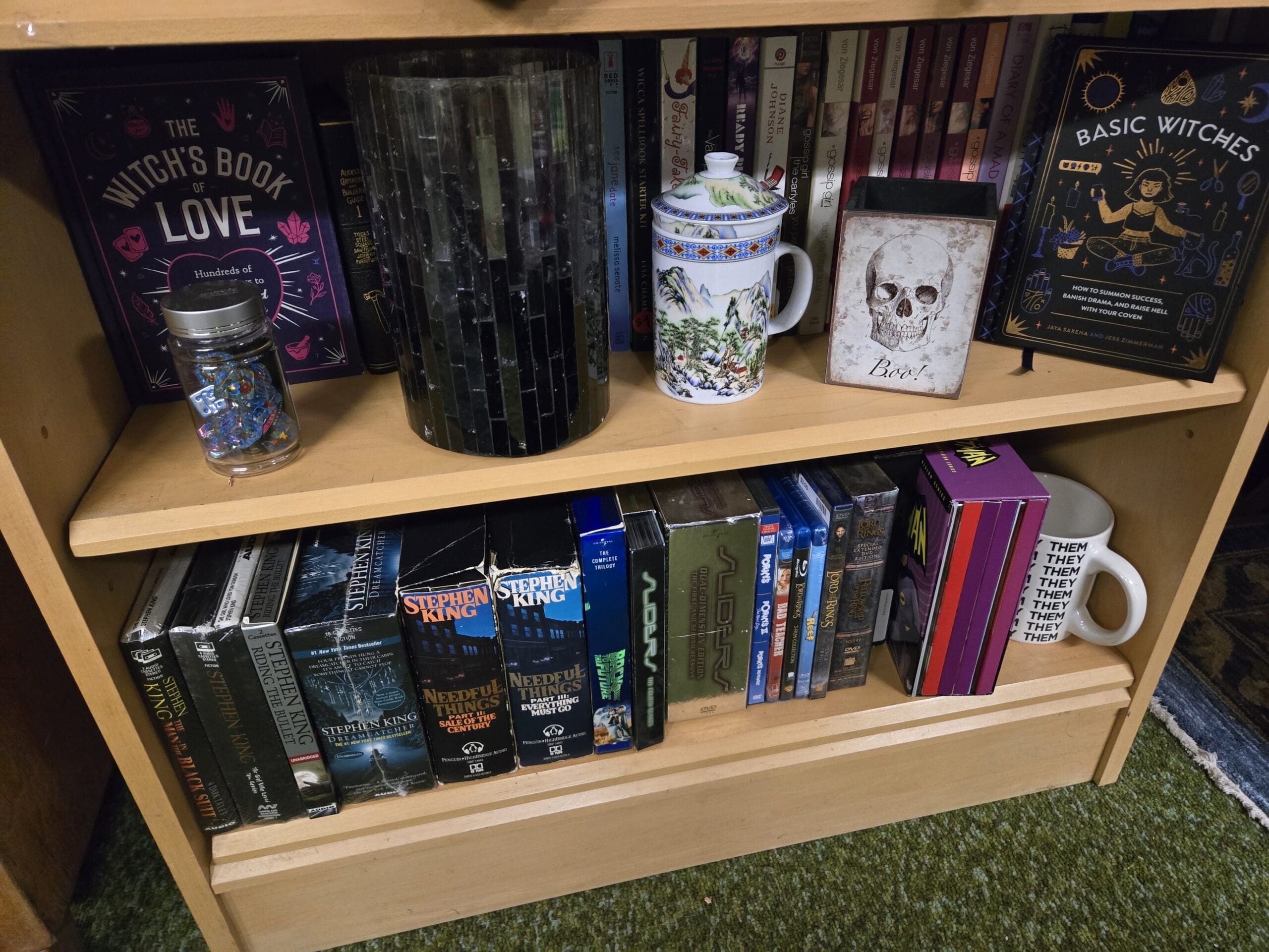 Bookshelf with The Witch's Book of Love, Basic Witches by Jaya Saxena and Jess Zimmerman, a Diane Johnson title, a Wicca spell book starter kit, an Asian-style porcelain tea mug with mountain landscape and beaded handle, a Boo Halloween skull frame, a candle holder, and a glittery confetti jar on the upper shelf, with Stephen King Needful Things Part 1 and Part 2 paperbacks, Stephen King Dreamcatcher and Riding the Bullet, a Sliders Complete Trilogy VHS set, a Lord of the Rings extended DVD set, and a THEM mug on the lower shelf at Shop 75, Pacific Antiques Mall in Tacoma, WA.