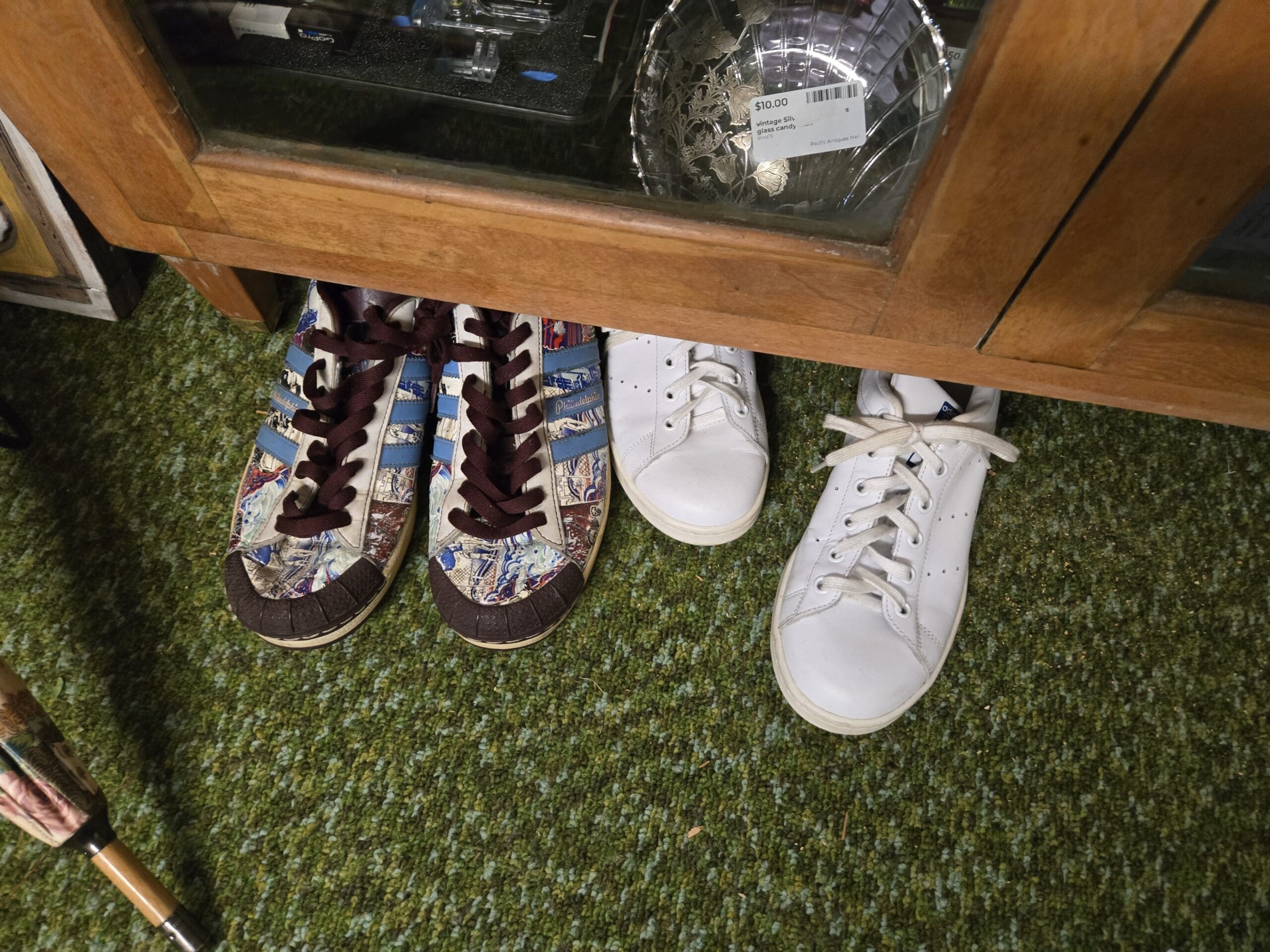 Three sneakers on the green carpet under a glass cabinet, including a pair of patterned Adidas Superstar-style sneakers with newspaper-style print, blue trefoil stripes, and brown shell toes and laces, and a pair of plain white leather Stan Smith-style tennis shoes, with a vintage silver-overlay etched glass candy dish visible in the cabinet above at Shop 75, Pacific Antiques Mall in Tacoma, WA.