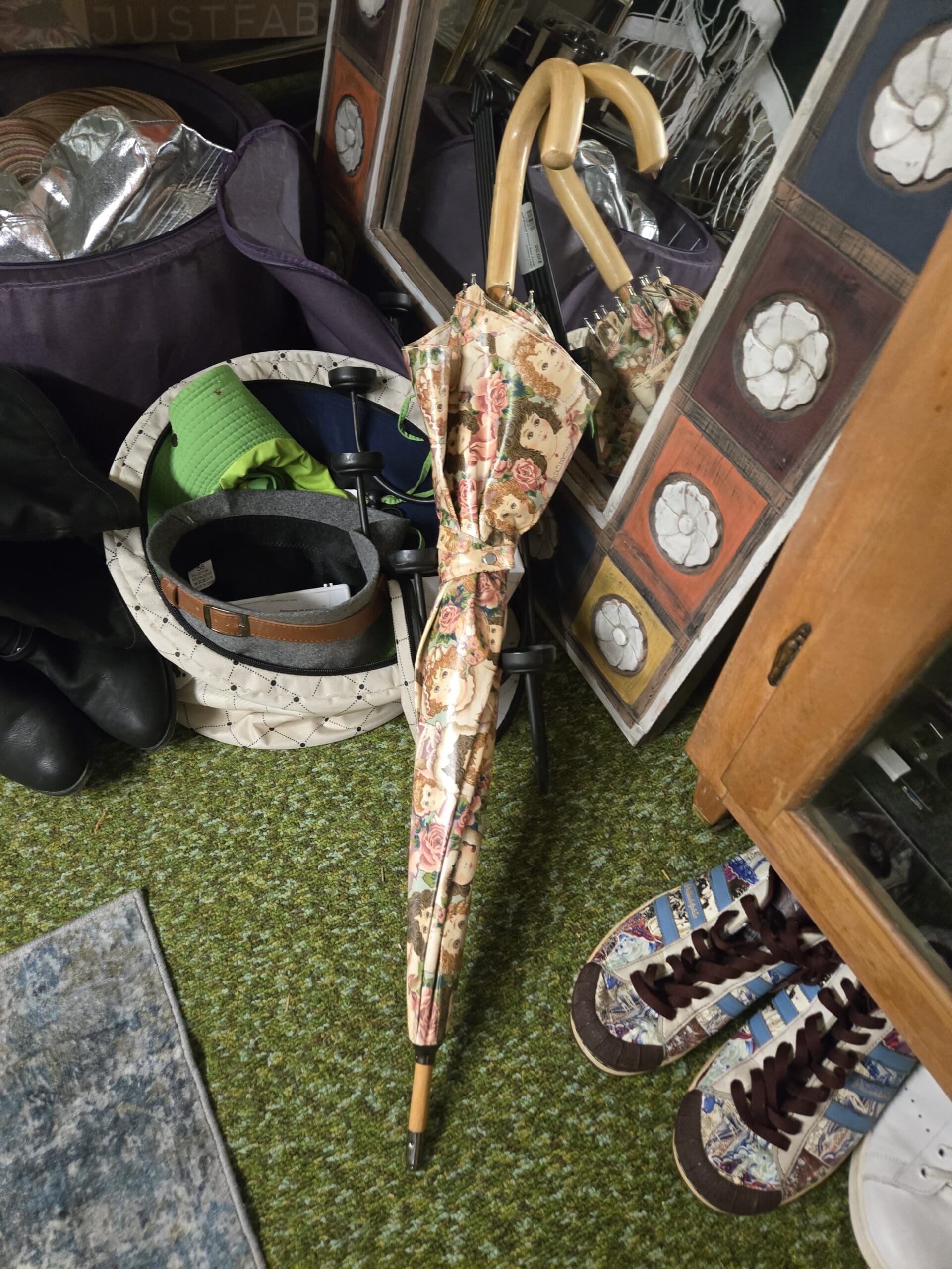 A vintage stick umbrella with cream cherubs-and-roses print canopy, curved wooden cane handle, and brass tip, leaning against a quilted cream tote bag holding green and white visors, with a JustFab boot box, black knee-high leather boots, and the floral tile-bordered standing mirror in the background, and patterned Adidas Superstar style sneakers visible on the right at Shop 75, Pacific Antiques Mall in Tacoma, WA.