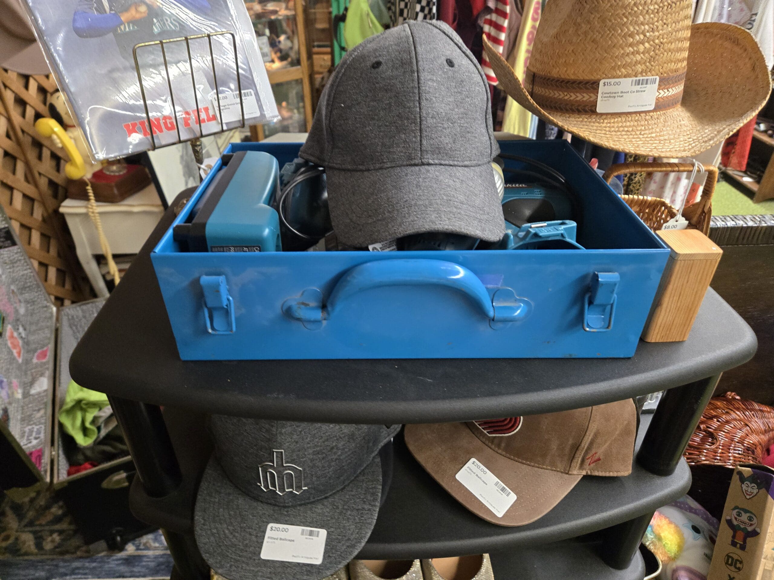 Display with a blue metal industrial divided toolbox on top holding a charcoal heather baseball cap and additional headwear, a Cowtown Boot Co straw cowboy hat to the right, a King Felix Seattle Mariners framed item on the left, and a lower shelf holding a gray Seattle Mariners fitted ball cap and a tan brown Cardinals style fitted ball cap at Shop 75, Pacific Antiques Mall in Tacoma, WA.