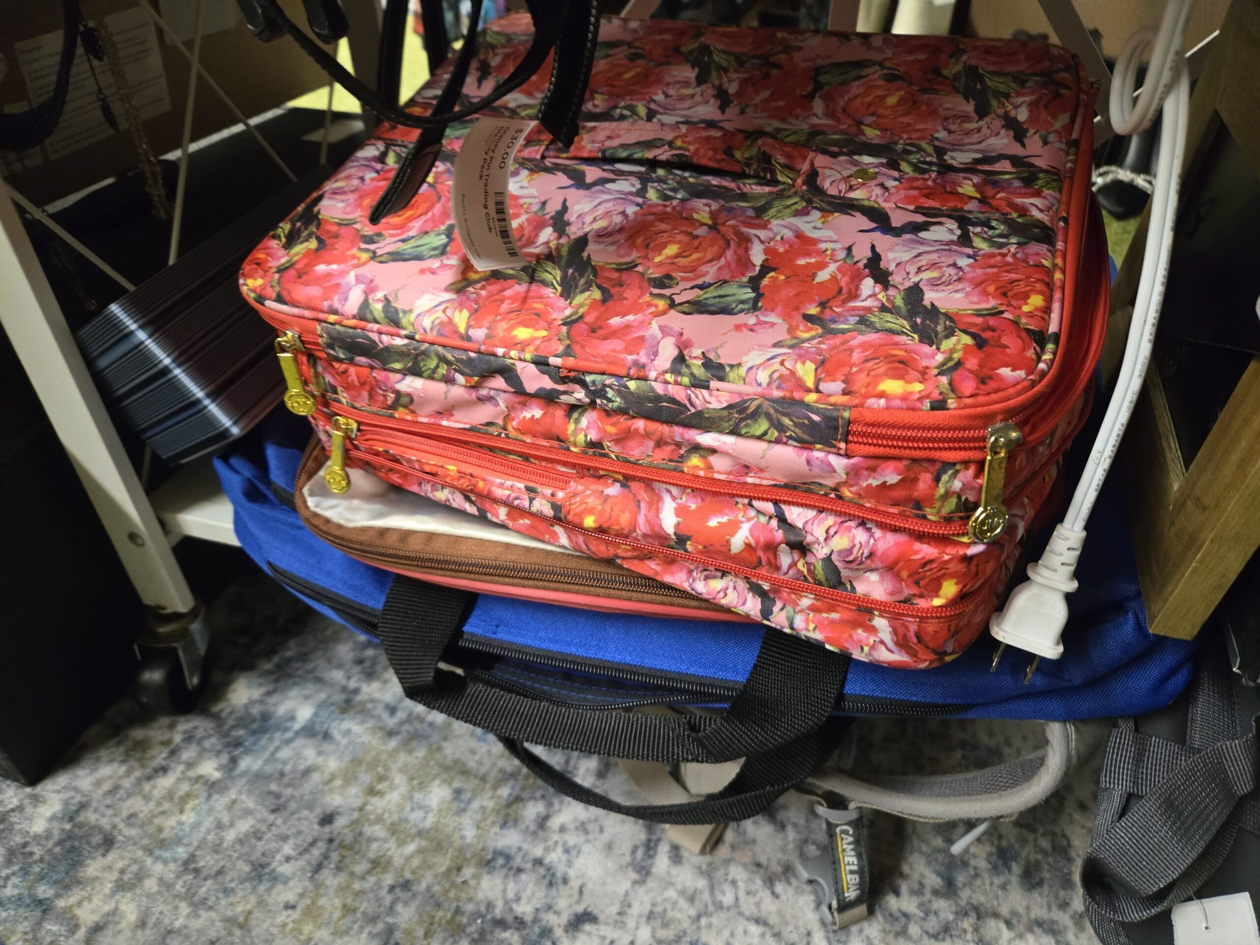 Stacked luggage with a soft red and pink rose printed makeup or laptop case with gold zipper pulls on top, a blue duffel laptop bag with black handles below, and a CamelBak hydration pack visible to the right on a blue-gray patterned rug at Shop 75, Pacific Antiques Mall in Tacoma, WA.
