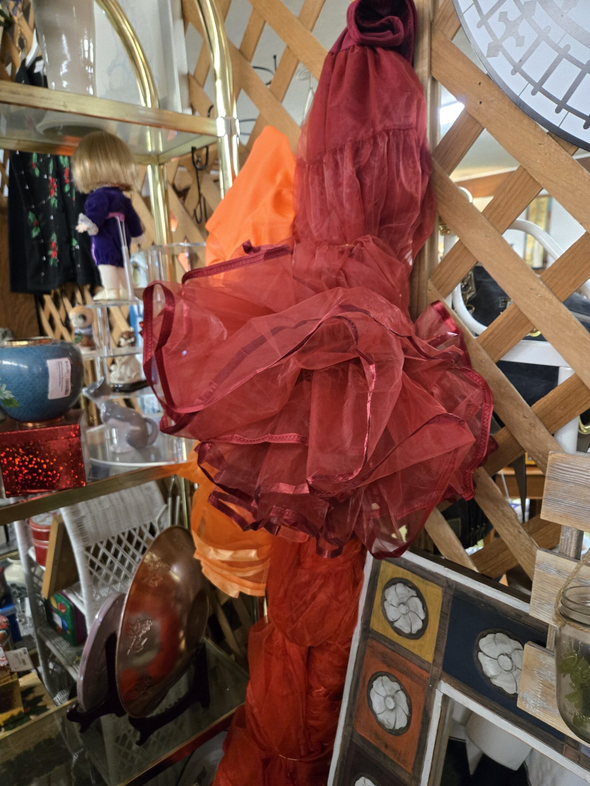 Sheer chiffon ruffled wraps in deep wine red and bright orange layered on the wood lattice wall, with a vintage porcelain doll in a purple and red Christmas-print dress visible on the brass shelf to the left, a small navy ceramic vase, a red glittery rectangular box, and a bronze decorative plate with rose relief at Shop 75, Pacific Antiques Mall in Tacoma, WA.