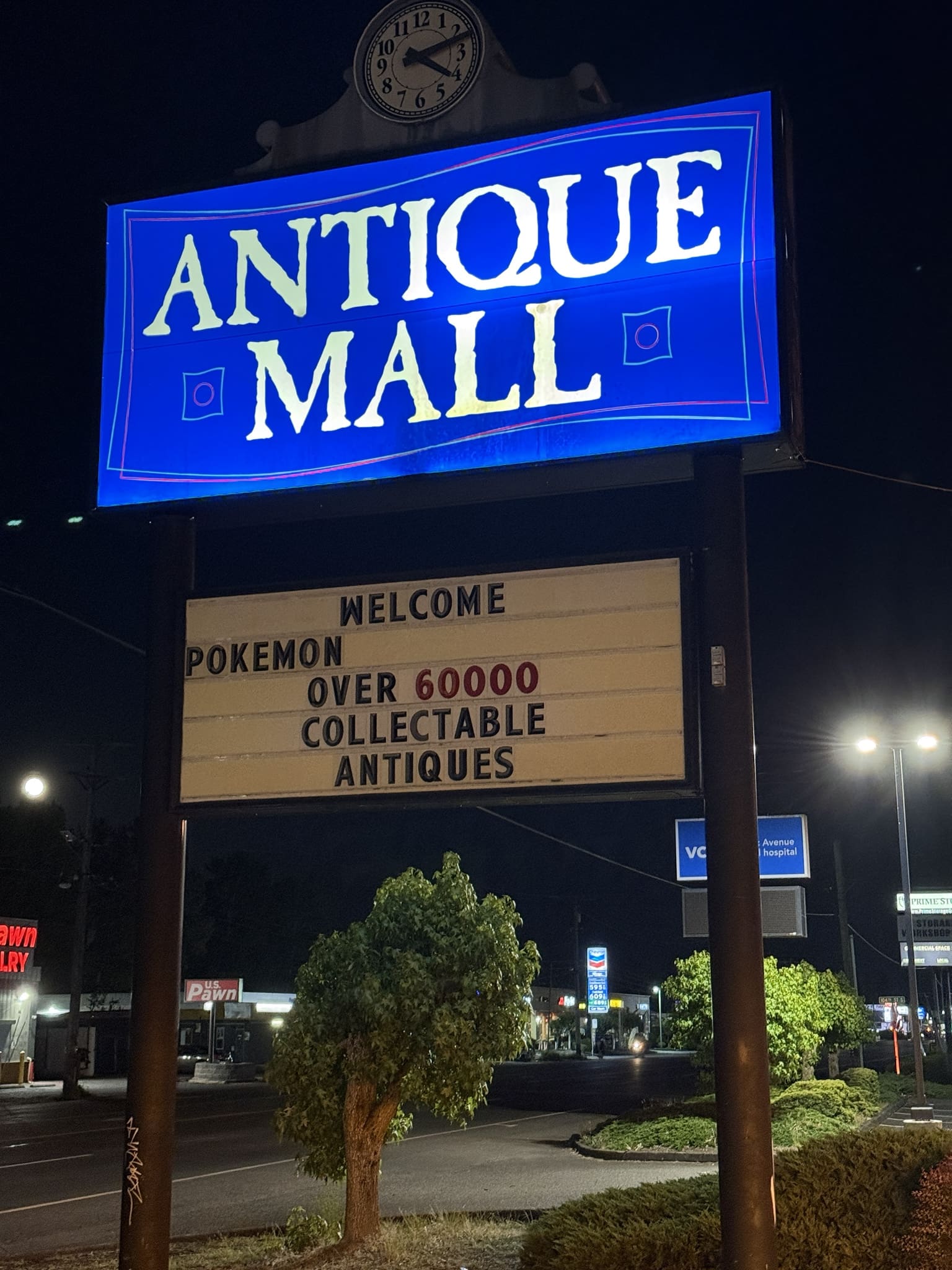 Nighttime view of the re-lit Pacific Antiques Mall pylon sign, with the ANTIQUE MALL panel glowing brilliant blue from internal lighting, the white lettering and pink-coral decorative dots illuminated, the Arabic-numeral clock above, and the letter-board marquee below reading WELCOME POKEMON OVER 60000 COLLECTABLE ANTIQUES, with US Pawn Jewelry, Chevron, and other Pacific Avenue businesses lit in the background at Pacific Antiques Mall in Tacoma, WA.