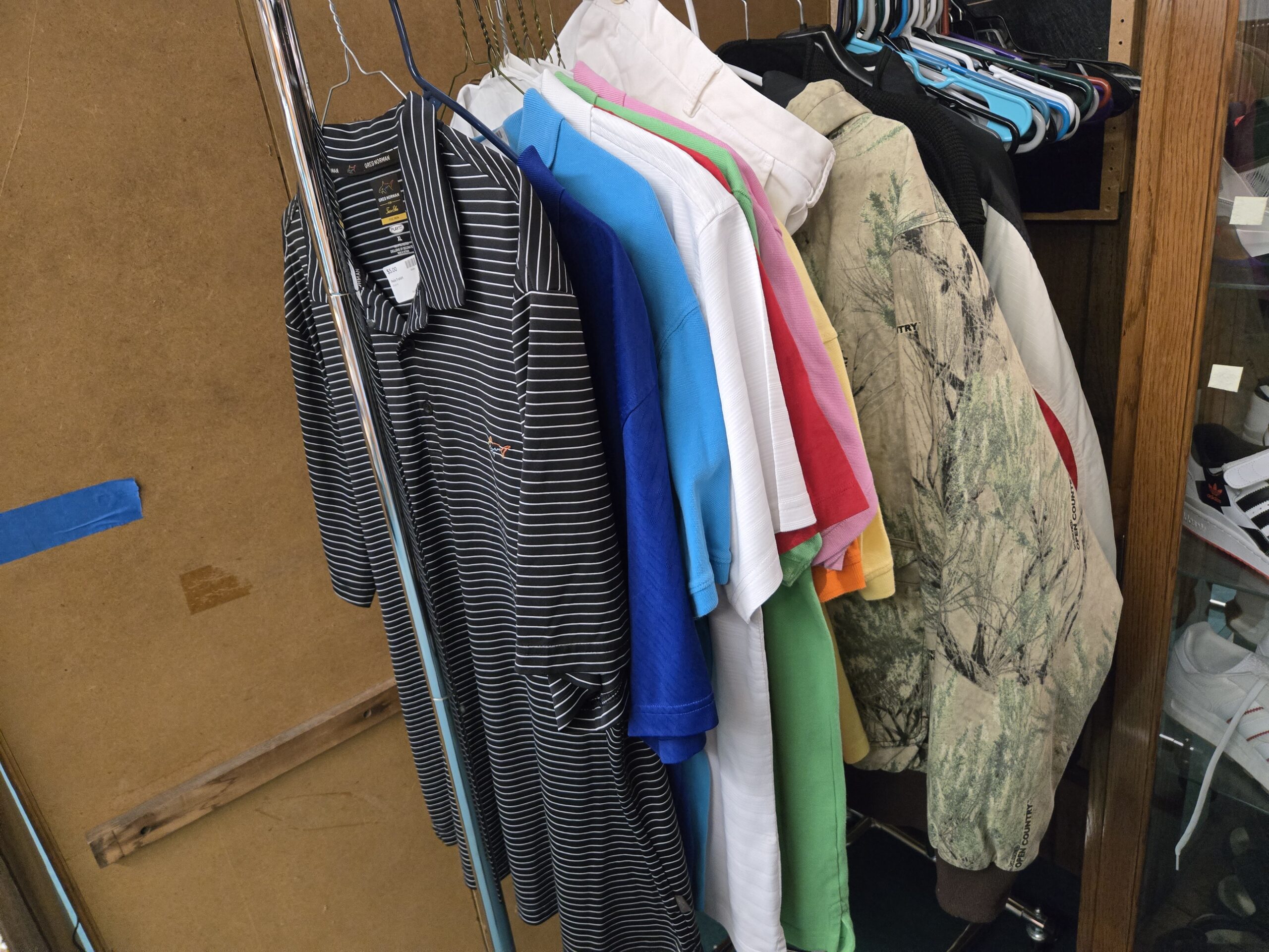Greg Norman black and white striped golf polos in XL, short-sleeve polos in royal blue, light blue, white, red, pink, green and yellow, white chinos, a Realtree Open Country camo hoodie in cream and sage with brown ribbed cuffs, and a black waffle-knit hoodie on a chrome rack next to an oak cabinet holding adidas three-stripe sneakers and white trainers at Shop 40, Pacific Antiques Mall in Tacoma, WA.