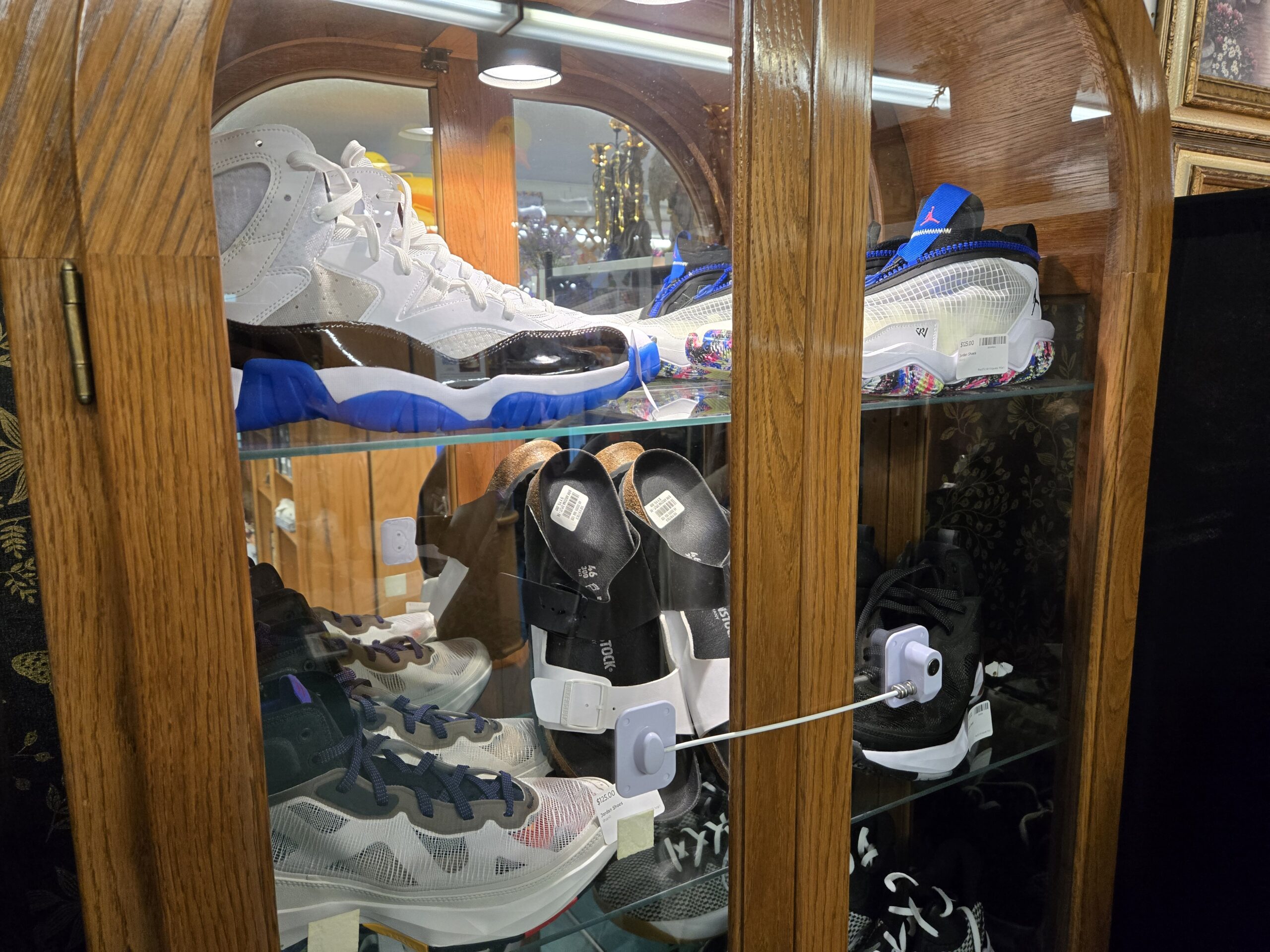 White and royal blue patent-leather Jordan mid-tops, a white and stone-grey Jordan high-top, a white Jordan pair with a royal blue ankle strap over a confetti-speckled sole, grey and purple laced Jordan mids, black knit Jordan high-tops, and black Birkenstock sandals with cork-lined footbeds on the glass shelves of a lit arched oak cabinet at Shop 40, Pacific Antiques Mall in Tacoma, WA.