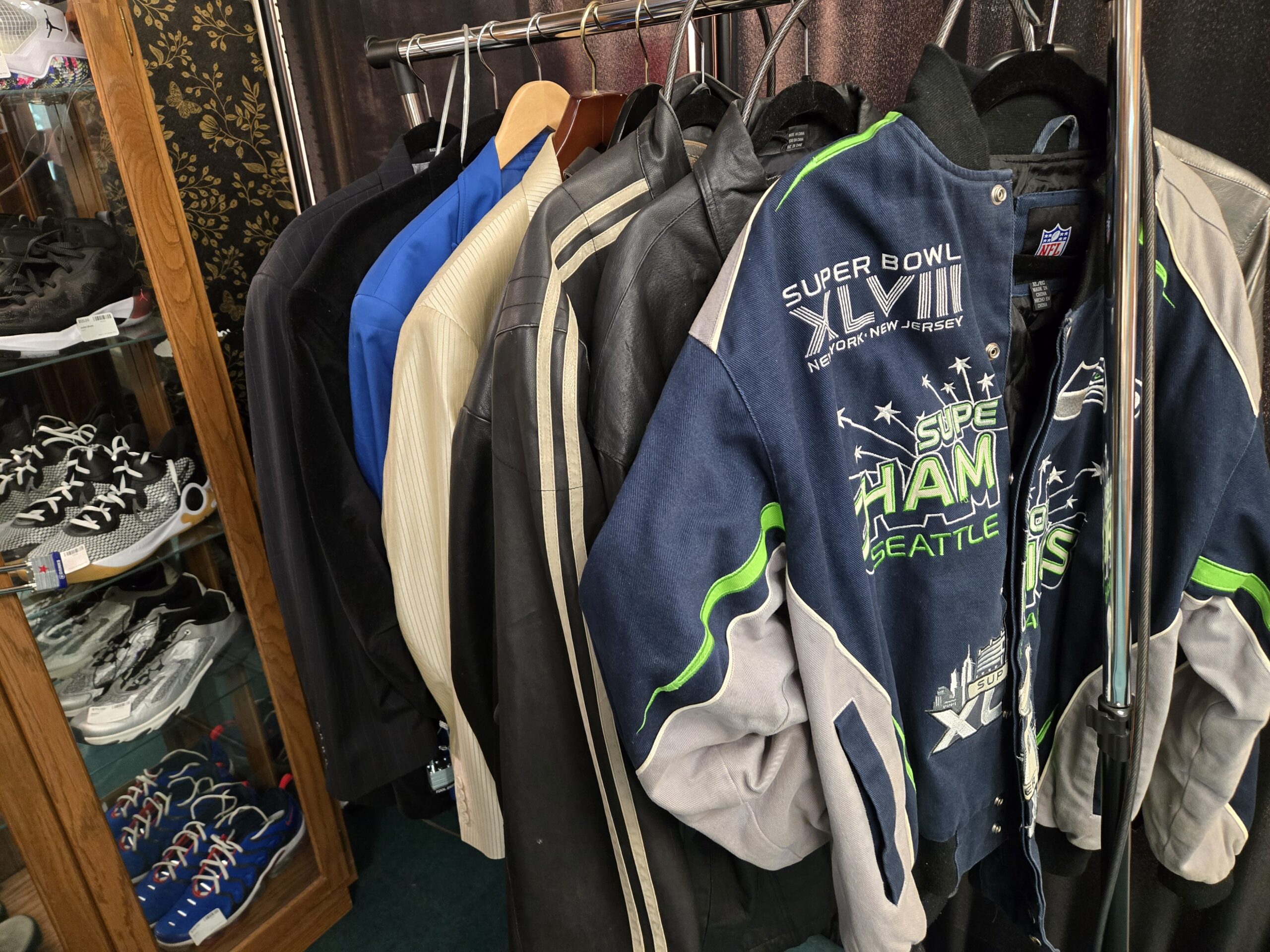 A Seattle Seahawks Super Bowl XLVIII jacket in navy, grey and green with New York, New Jersey embroidery and NFL labels in XL, a black leather jacket with cream racing stripes, an ivory ribbed-panel leather jacket, a royal blue jacket, and a black pinstripe sport coat on a chrome rack beside an oak cabinet holding black and white knit high-tops, grey mesh sneakers, and royal blue high-tops at Shop 40, Pacific Antiques Mall in Tacoma, WA.