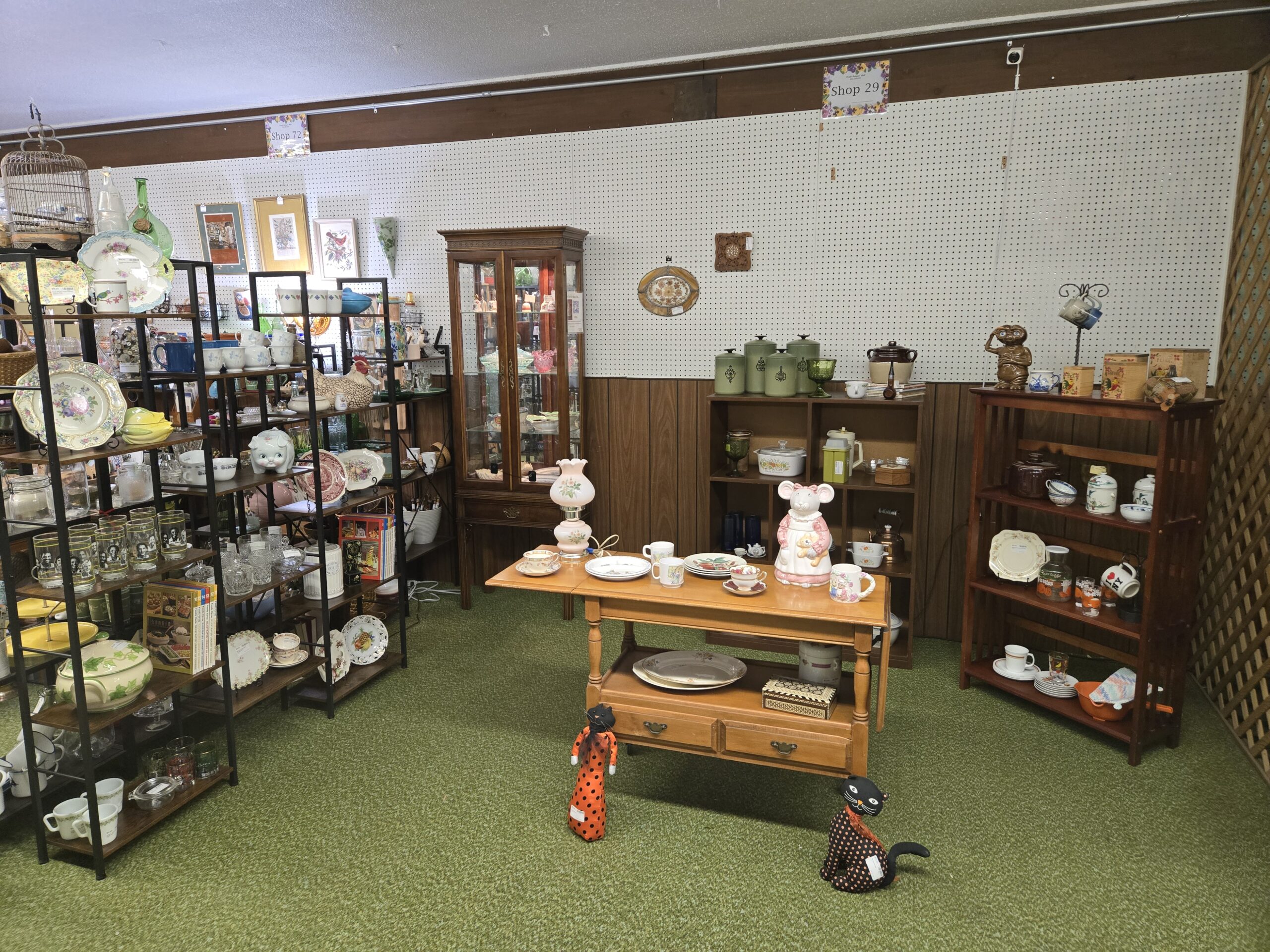 Wide view of the Shop 29 space under its pegboard sign, with black metal shelving holding floral dinnerware, pressed glass, Seattle SuperSonics tumblers, Family Circle cooking volumes, a floral piggy bank, and a ceramic hen on nest, a lighted walnut curio cabinet, a drop-leaf table set with children's dishes and a mouse cookie jar, cube shelving with avocado green canisters, a wooden shelf holding an E.T. cookie jar and a rooster canister set, and two polka-dot Halloween cats on green carpet at Shop 29, Pacific Antiques Mall in Tacoma, WA.