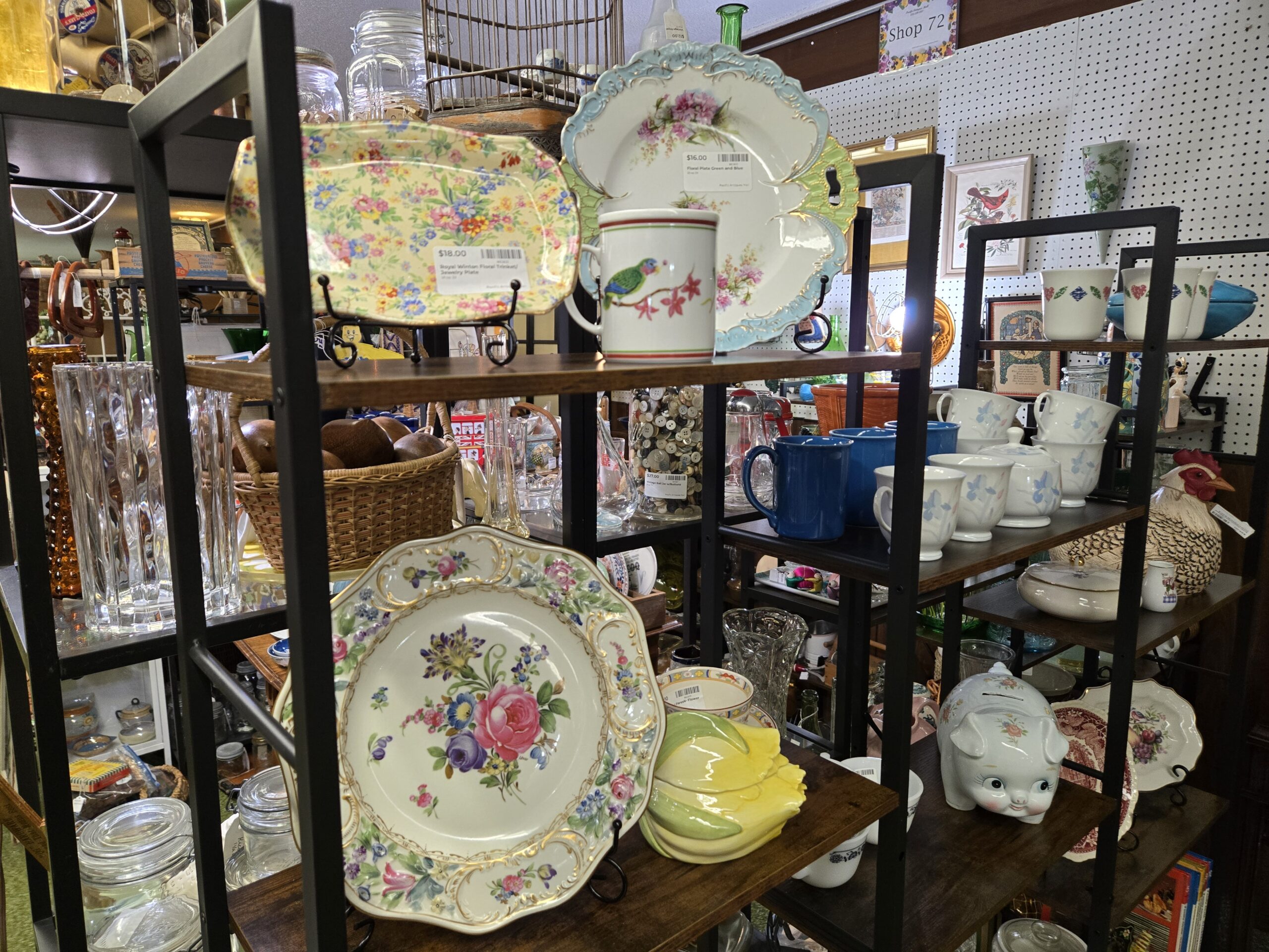 A Royal Winton floral chintz trinket plate on a yellow ground, an embossed green and blue plate with pink blossoms, a mug painted with a parrot and red flowers, a large plate centered on a pink rose with gold scrollwork, a yellow corn-form dish, blue stoneware mugs, white mugs with blue iris relief and a matching lidded sugar bowl, a speckled ceramic hen on nest, a floral piggy bank, a glass jar packed with old buttons, amber and clear cut glass vases, and framed cardinal and botanical prints at Shop 29, Pacific Antiques Mall in Tacoma, WA.