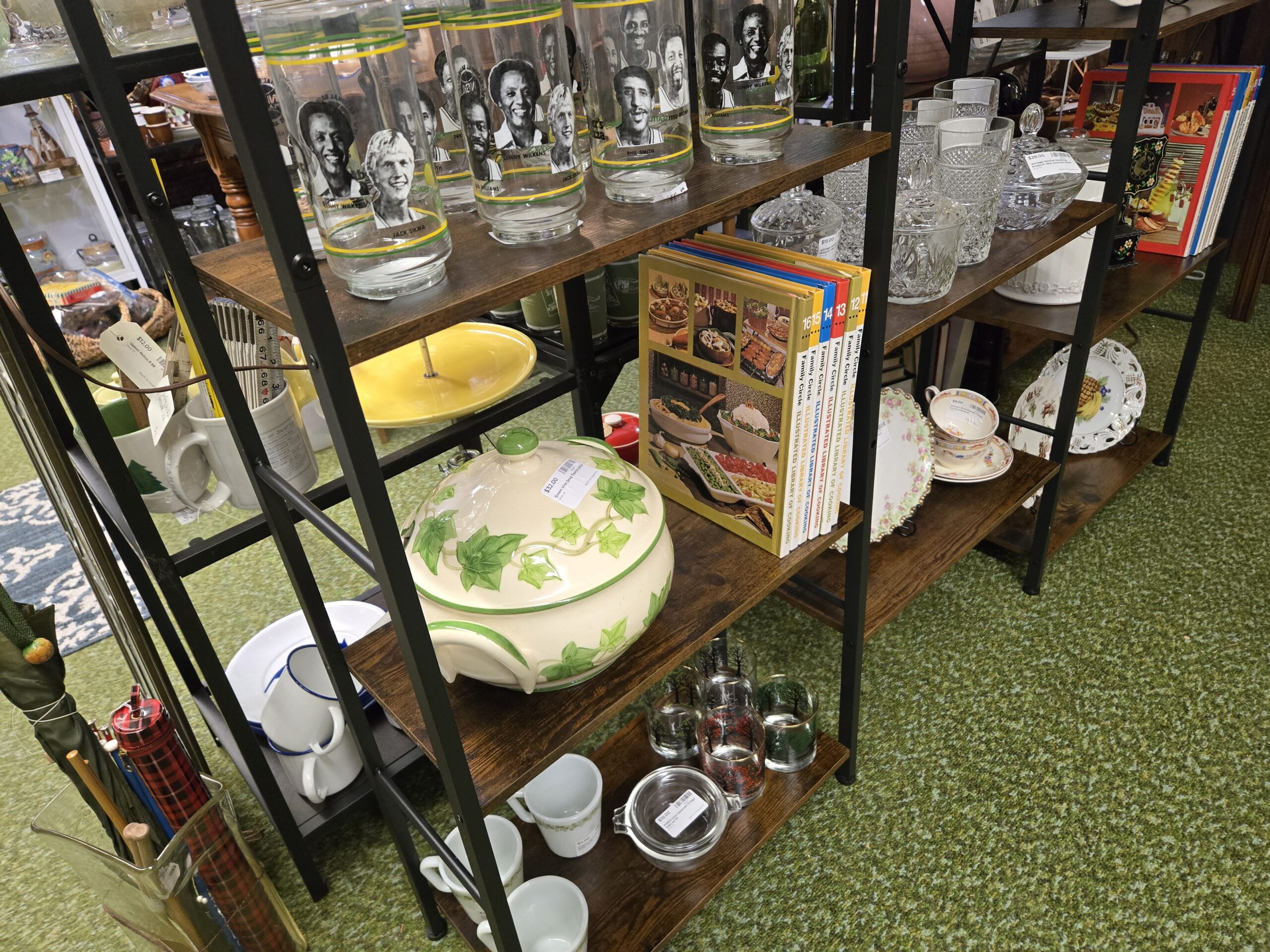 Green and gold Seattle SuperSonics player tumblers naming Jack Sikma, Lenny Wilkens, Gus Williams, Fred Brown, and Phil Smith, a cream and green ivy-vine covered tureen, Family Circle Illustrated Library of Cooking volumes eleven through sixteen, Anchor Hocking Prescut lidded candy dishes with diamond-pattern tumblers, a Limoges plate with pink roses and a gold scalloped rim, a floral teacup and saucer, rocks glasses painted with trees, and plaid umbrellas in a stand at Shop 29, Pacific Antiques Mall in Tacoma, WA.