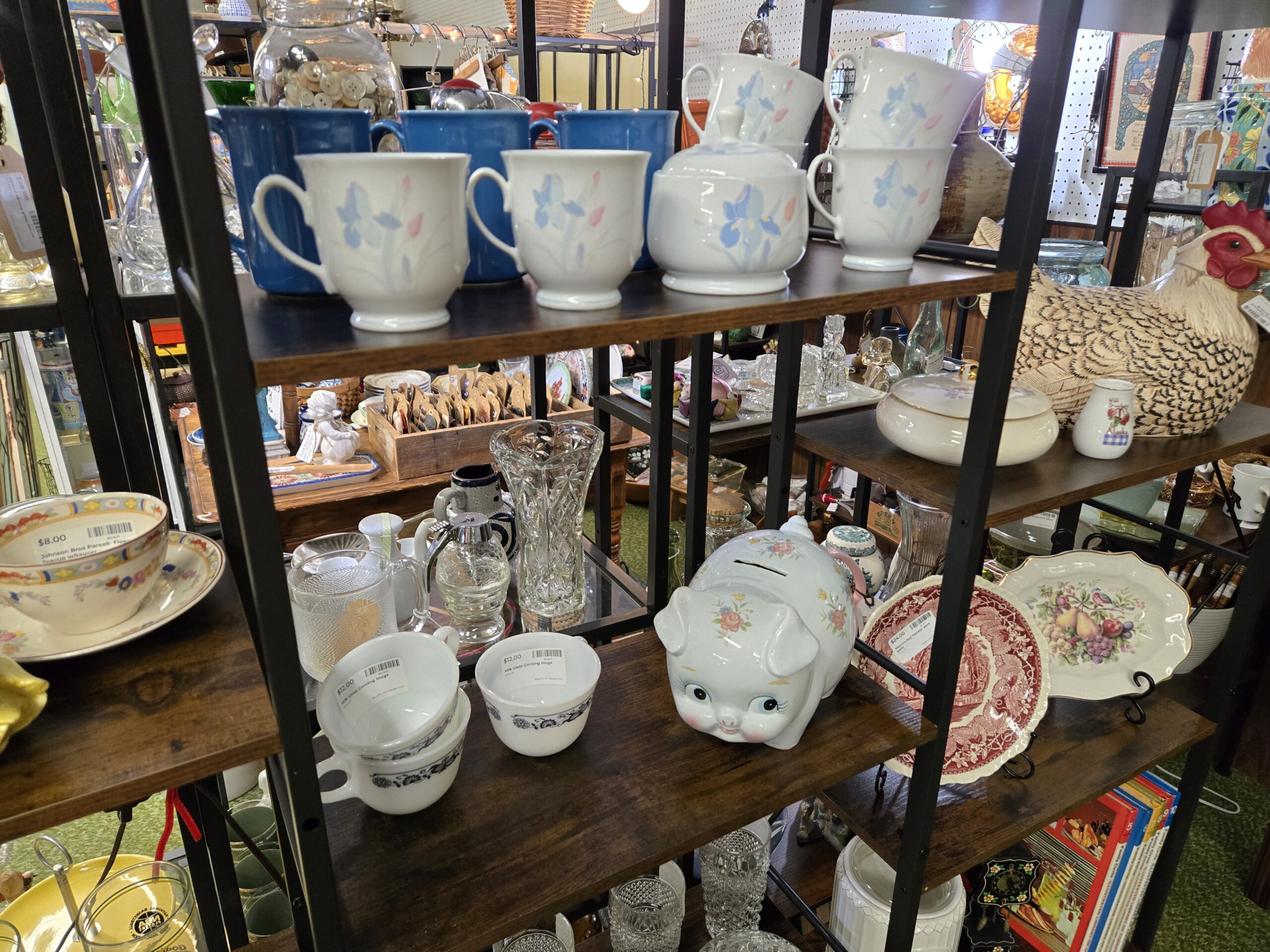 White mugs with blue iris relief and a matching lidded sugar bowl, blue stoneware mugs, milk glass Corning mugs in a blue and white floral pattern, a floral piggy bank, a Mason's red transferware plate, a platter decorated with fruit and a bluebird under a gold rim, a Johnson Bros Pareek teacup and saucer with a multicolored border, a cut glass vase, a glass syrup pitcher, a jar of white buttons, a speckled ceramic hen on nest, and a small Wild Strawberry Alaska pitcher at Shop 29, Pacific Antiques Mall in Tacoma, WA.