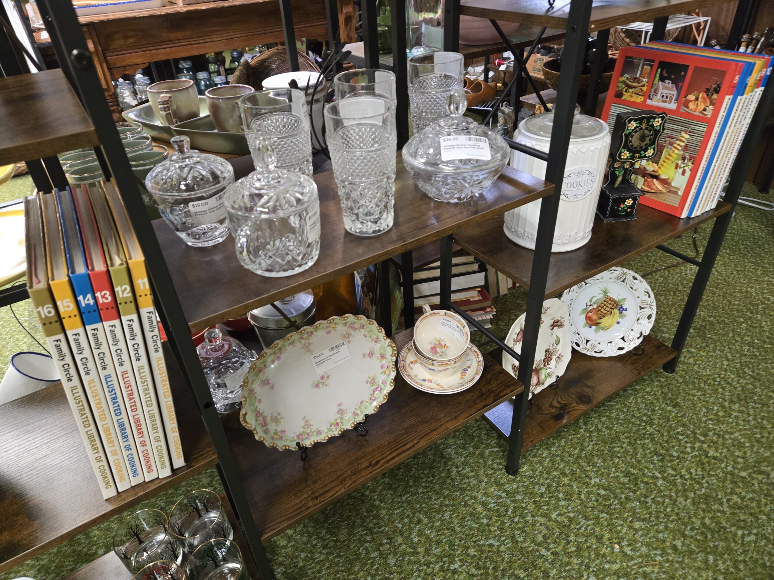 Clear pressed glass including Anchor Hocking Prescut lidded candy dishes in the Star and Fan pattern, a footed bowl, and tall diamond-pattern tumblers, a white ribbed ceramic cookie jar, a black painted wall clock with floral decoration, Family Circle Illustrated Library of Cooking volumes eleven through sixteen, a Blakeman and Henderson Limoges platter with pink roses and a gold scalloped rim, a floral teacup and saucer, a fruit-decorated square plate, and a pierced-rim plate with pineapple and grapes at Shop 29, Pacific Antiques Mall in Tacoma, WA.