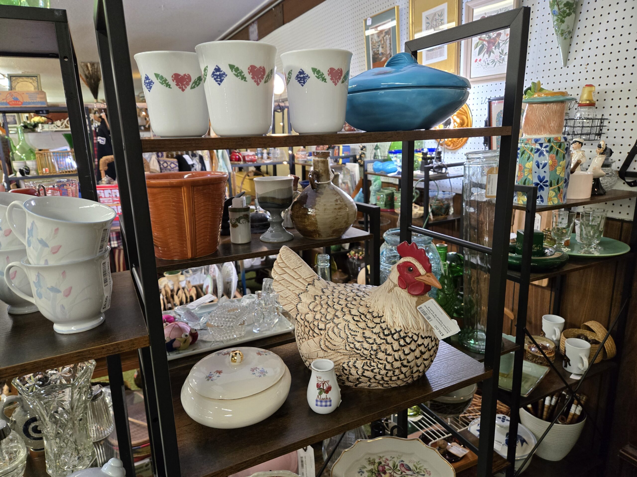 Three white cups with folk-art heart and leaf motifs, a teal lidded casserole, a terracotta basketweave planter, a gray and brown stoneware goblet, a brown drip-glaze jug, a speckled ceramic hen on nest with a red comb, a cream lidded dish with a gold knob and floral sprays, a small Wild Strawberry Alaska pitcher, white cups with blue and pink iris relief, and framed botanical prints on the pegboard at Shop 29, Pacific Antiques Mall in Tacoma, WA.