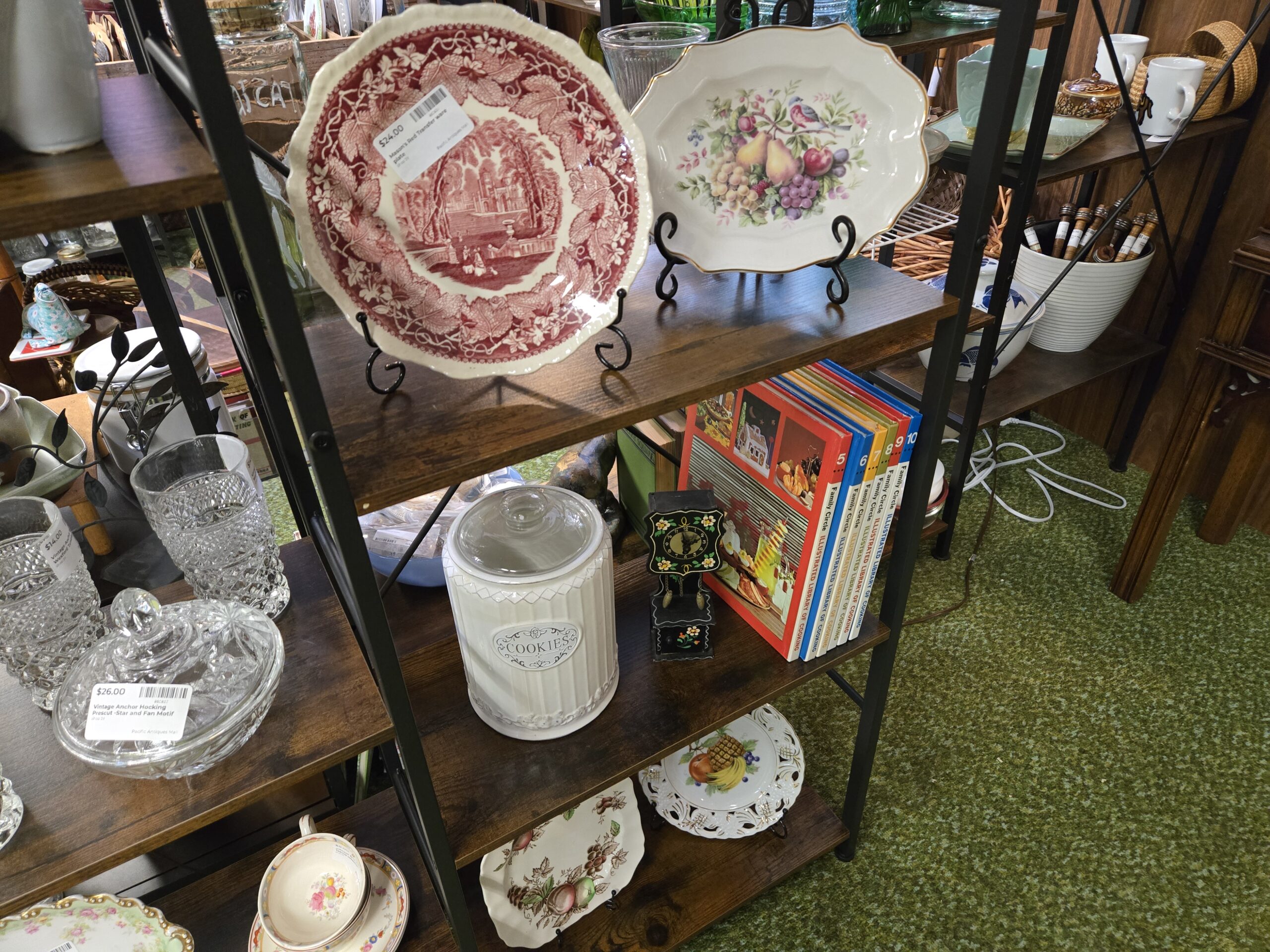 A Mason's red transferware plate with a castle scene on a wire stand, an oval platter decorated with fruit and a bluebird under a gold rim, a white ribbed ceramic cookie jar, a small black painted clock with floral trim, Family Circle Illustrated Library of Cooking volumes five through ten, Anchor Hocking Prescut pressed glass with diamond-pattern tumblers and a lidded bowl, a floral teacup and saucer, and a pierced-rim fruit plate at Shop 29, Pacific Antiques Mall in Tacoma, WA.