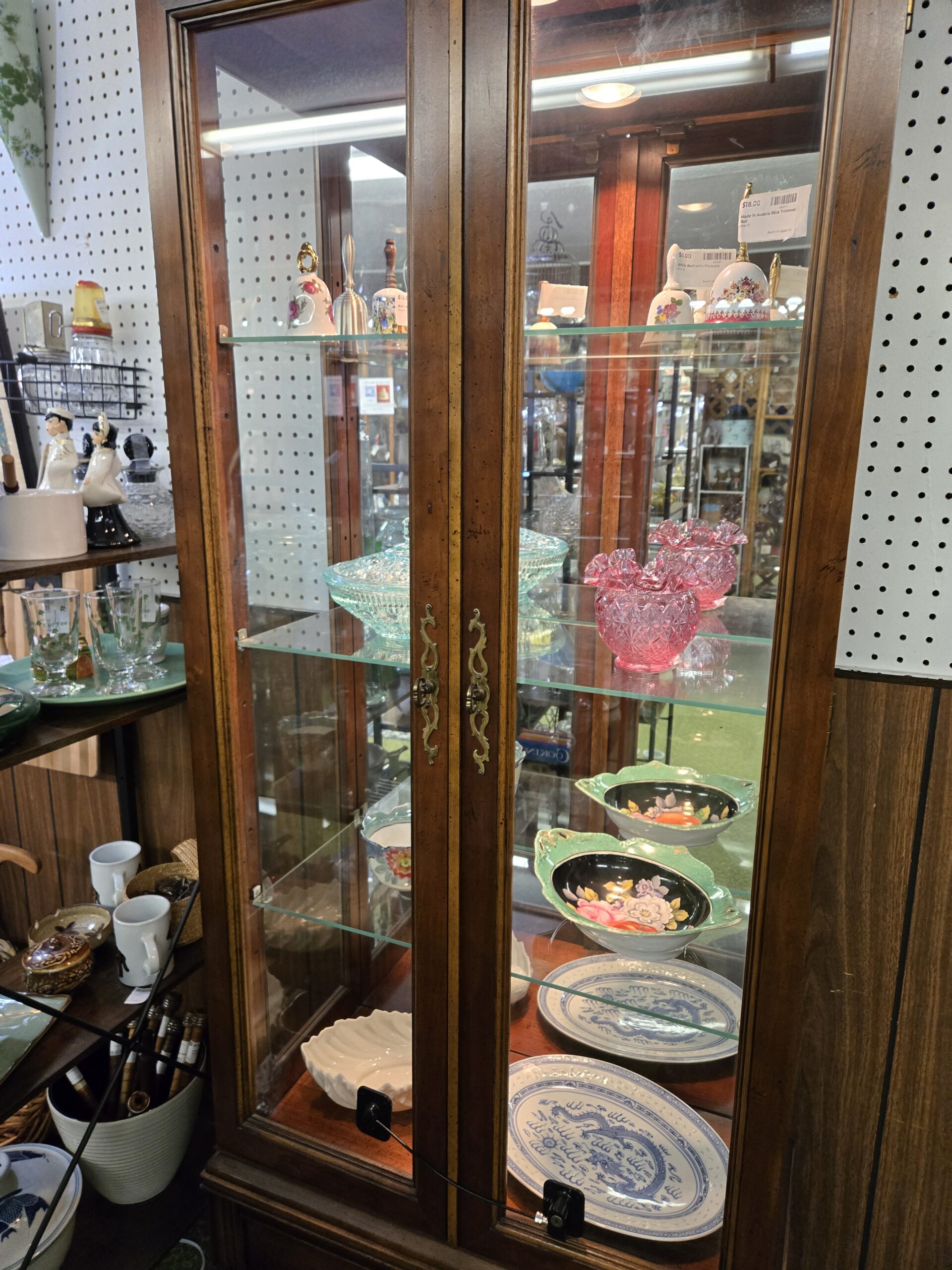 A lighted walnut curio cabinet with brass handles holding porcelain and metal bells including a pink-trimmed Austrian bell and a white bell with painted flowers, an aqua pressed glass footed bowl, pink ruffled pressed glass vases in a diamond pattern, hand-painted bowls with mint green scalloped rims and floral centers on black grounds, blue and white dragon-pattern plates, and a white scalloped shell dish at Shop 29, Pacific Antiques Mall in Tacoma, WA.