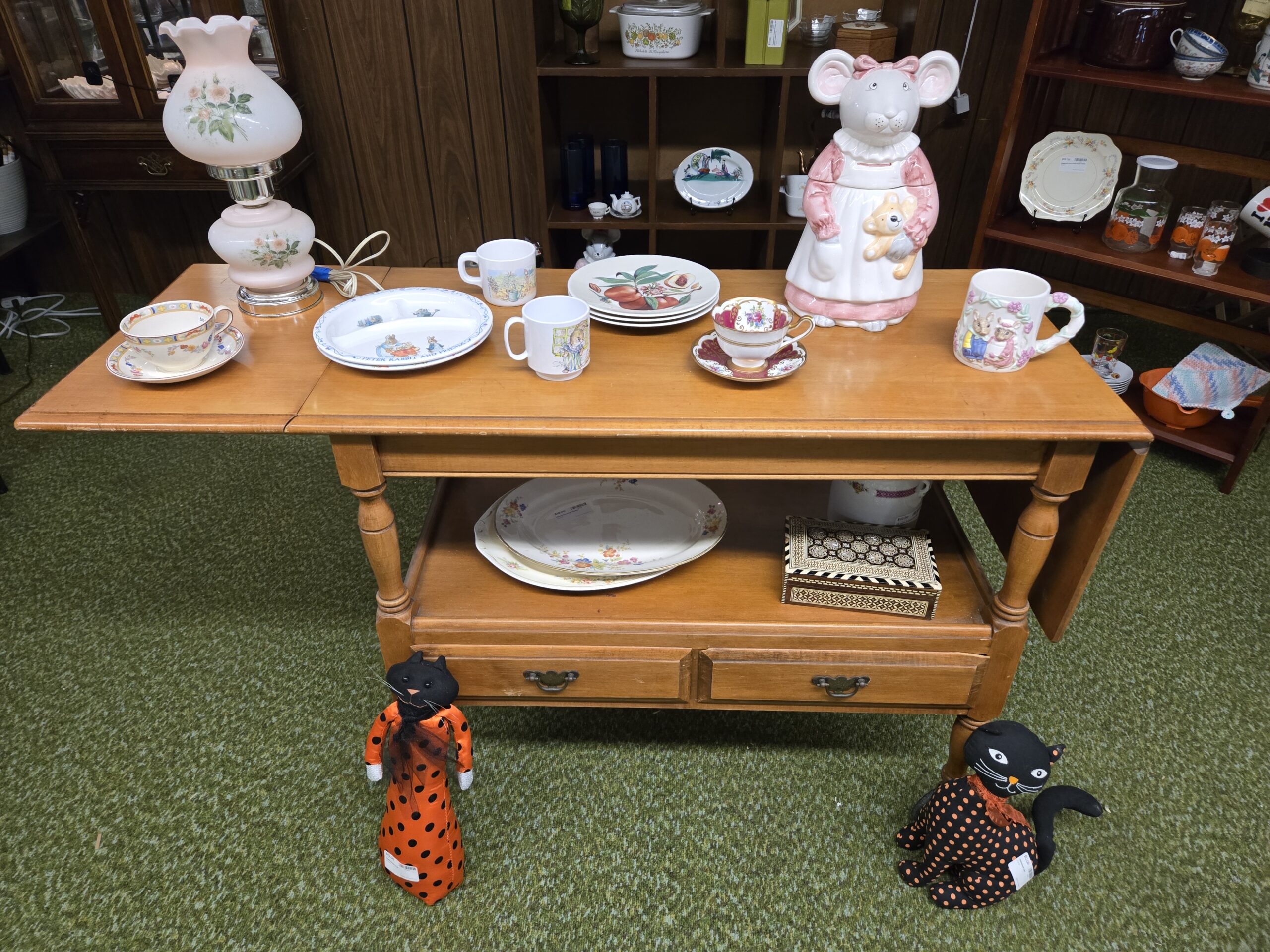 A drop-leaf table set with a Peter Rabbit and Friends divided child's plate, two children's mugs with rabbit scenes, a relief-molded rabbit mug with a vine handle, a pink glass hurricane lamp painted with roses, a mouse cookie jar in a pink apron holding a teddy bear, a gold and burgundy teacup and saucer, stacked botanical fruit plates, an inlaid marquetry box, floral dinner plates on the lower shelf, and two orange and black polka-dot fabric Halloween cats at Shop 29, Pacific Antiques Mall in Tacoma, WA.