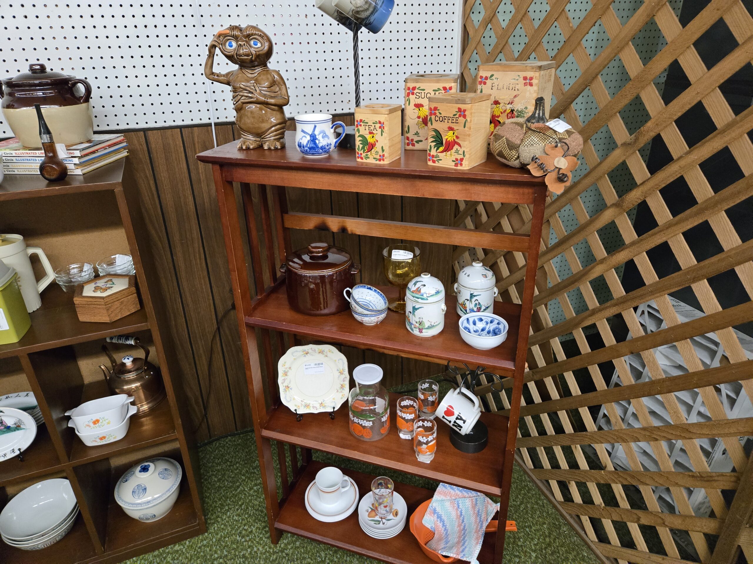 A brown-glazed E.T. figural cookie jar, a Delft blue windmill mug, a four-piece wooden canister set with rooster decals marked tea, coffee, sugar, and flour, a patchwork fabric pumpkin, a brown and cream stoneware bean pot with lid, an amber pressed glass goblet, hand-painted Asian lidded jars with figural scenes, blue and white rice-pattern bowls, a floral scalloped dinner plate, an orange citrus juice carafe with matching tumblers, an I Love NY mug, a copper kettle, and a Corning Ware petite pan with orange florals at Shop 29, Pacific Antiques Mall in Tacoma, WA.