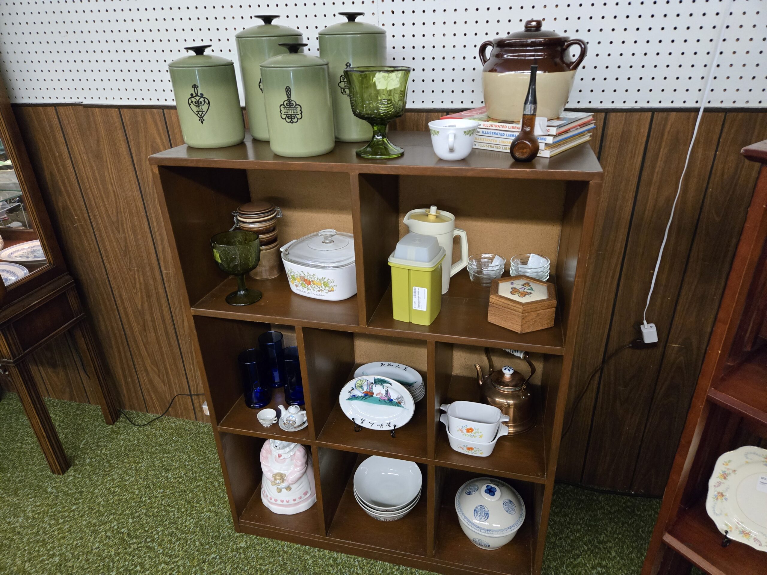 An avocado green kitchen canister set with black knobs marked tea, coffee, and sugar, green pressed glass goblets, a brown and cream stoneware bean pot with lid, a Corning Ware casserole with its glass lid, cobalt blue tumblers, glass custard cups, an oak hexagonal box with a butterfly under glass, a copper kettle with a white porcelain handle, hand-painted platters with calligraphy and figural scenes, a blue and white lidded rice bowl, a mouse cookie jar, and stacked bowls in wooden cube shelving at Shop 29, Pacific Antiques Mall in Tacoma, WA.