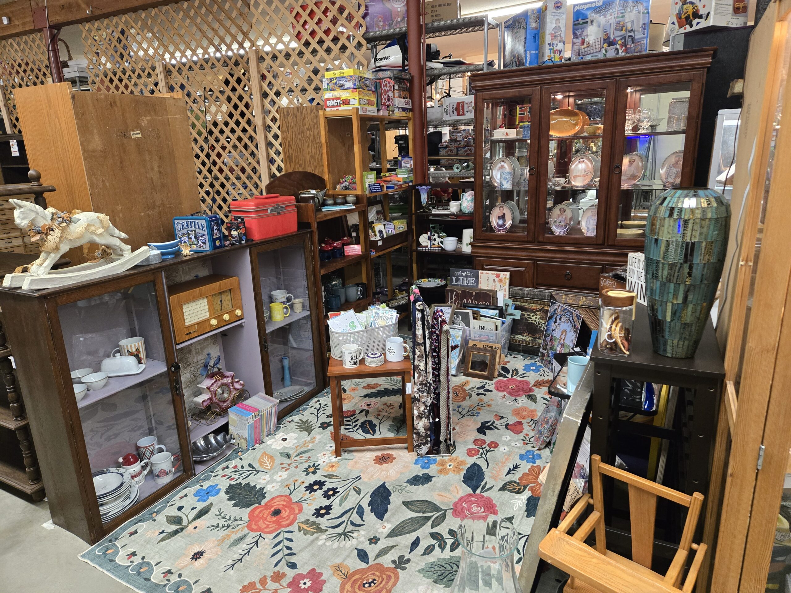 Wide view of Shop 116 with a papier-mache rocking horse trimmed in lace and paper flowers on an oak sliding-door cabinet, a blue Seattle Mariners tin lunchbox, a red vintage train case, party and trivia games stacked on pine shelving, a china cabinet of Princess Diana portrait collector plates, a tall teal mosaic tile floor vase, patterned neckties on a rack, greeting cards in a white basket, and painted wood wall signs on a floral area rug at Shop 116, Pacific Antiques Mall in Tacoma, WA.