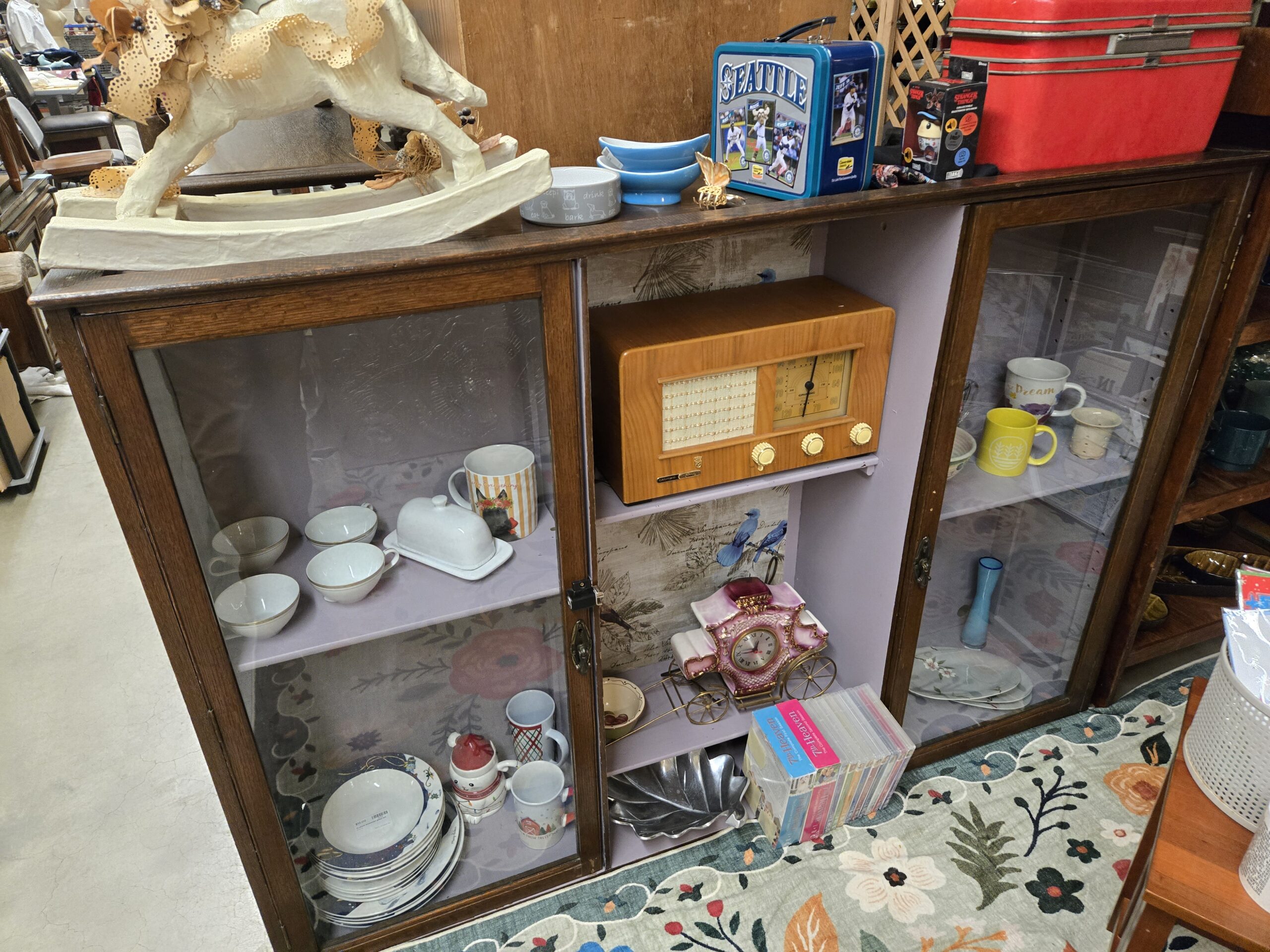 Oak cabinet with sliding glass doors holding white gold-rimmed teacups and bowls, a covered butter dish, stacked blue floral dinner plates, holiday mugs, and a yellow wheat-motif mug, with a Grundig wood-cased tabletop radio with gold dial and cream knobs on the center shelf above a pink and white porcelain carriage clock with gilt scrollwork and spoked brass wheels, an aluminum leaf-shaped dish, boxed 7th Heaven CD sets, a blue glass bud vase, and a Seattle Mariners tin lunchbox, Stranger Things figure box and red train case on top at Shop 116, Pacific Antiques Mall in Tacoma, WA.