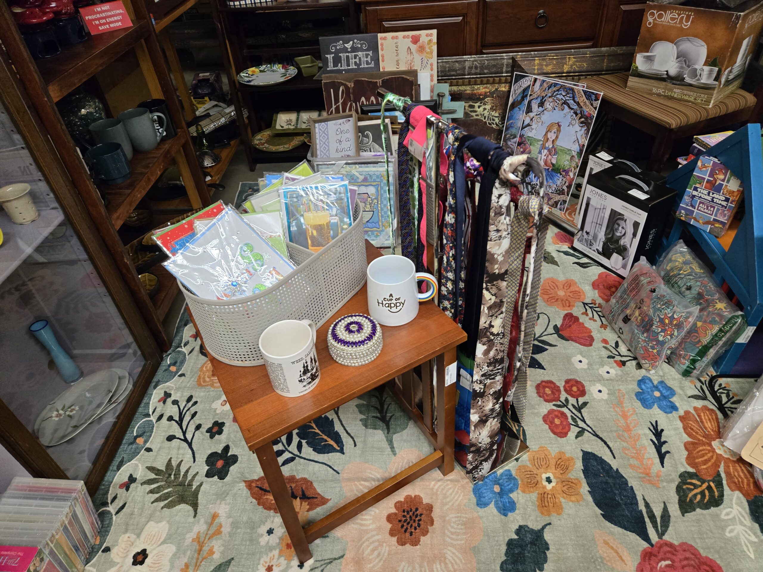 Teak side table holding a white perforated basket of sleeved greeting cards, a white A Cup of Happy mug, a Weed California souvenir mug, and a purple and white beaded lidded trinket box, with a rack of patterned neckties behind it, an Alice in Wonderland art print, Jones New York boxed tabletop sets, a Gallery Tabletops 16-piece dinnerware box, a blue painted wall shelf, a Land Before Time DVD collection, stoneware mugs on the shelf at left, and stacked CD cases at Shop 116, Pacific Antiques Mall in Tacoma, WA.