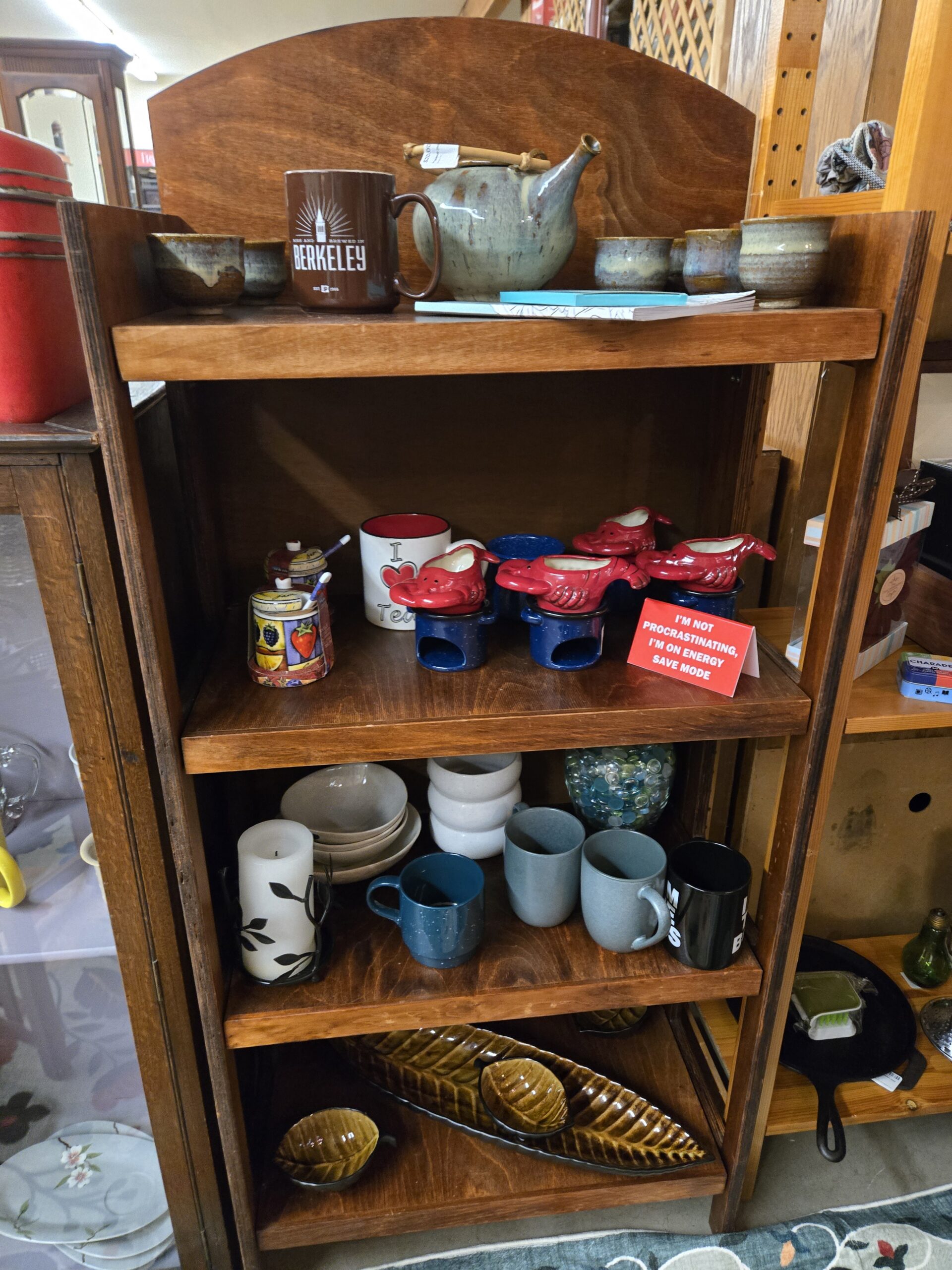 Stained wood shelf unit holding a speckled gray-green stoneware teapot with a bamboo handle, hand-thrown stoneware cups in drip glaze, and a brown Berkeley mug on top, red lobster-form ceramic lidded servers on blue speckled enamel warmers with a fruit-painted honey pot and dipper and an I Love Tea mug below, stacked stoneware bowls and plates with gray and blue matte mugs, a leaf-motif pillar candle in a metal holder and a jar of glass gems on the third shelf, and olive-glazed ceramic leaf-shaped serving dishes on the bottom at Shop 116, Pacific Antiques Mall in Tacoma, WA.