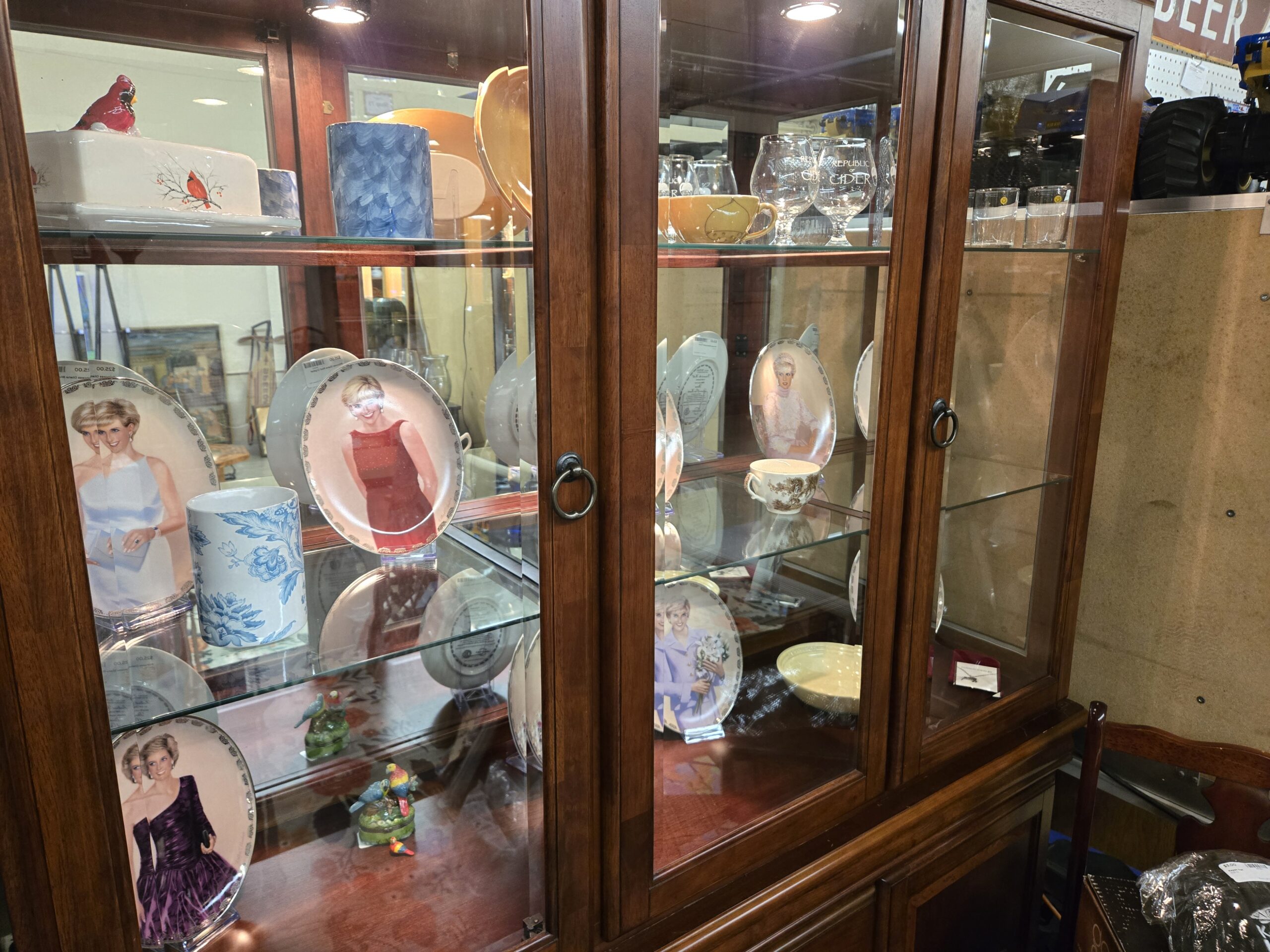 China cabinet holding Princess Diana portrait collector plates on stands with gilt scrollwork rims, showing Diana in a red beaded shift, a one-shoulder purple taffeta gown, a pale blue gown and a lilac suit holding white flowers, with additional plates turned to show the printed certificates on the reverse, a blue and white floral porcelain canister, a cardinal-decorated covered ceramic box, etched Republic of Cider tulip glasses, an amber teacup, a blue art glass vase and parrot and bluebird figural trinket boxes on mossy bases at Shop 116, Pacific Antiques Mall in Tacoma, WA.