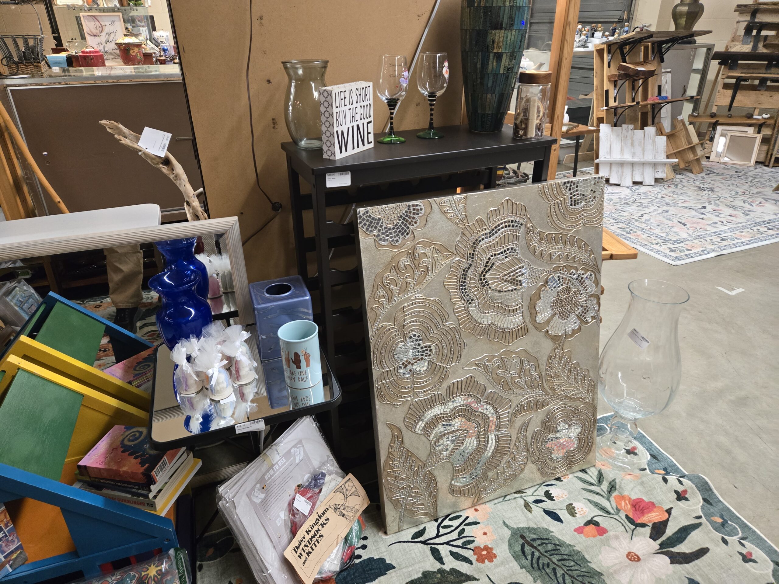Corner display with a taupe mosaic tile floral wall panel set with mirrored glass tesserae, clear glass hurricane vases, wine glasses with black and green striped stems, a Life Is Short Buy The Good Wine block sign, a cobalt blue glass vase, a blue ceramic tissue box cover, a We Are One Human Race mug, tulle-wrapped candle favors, a black metal wine rack console table, a tall teal mosaic tile floor vase, a jar of wine corks, a driftwood branch and packaged Daisy Kingdom windsocks and kites at Shop 116, Pacific Antiques Mall in Tacoma, WA.