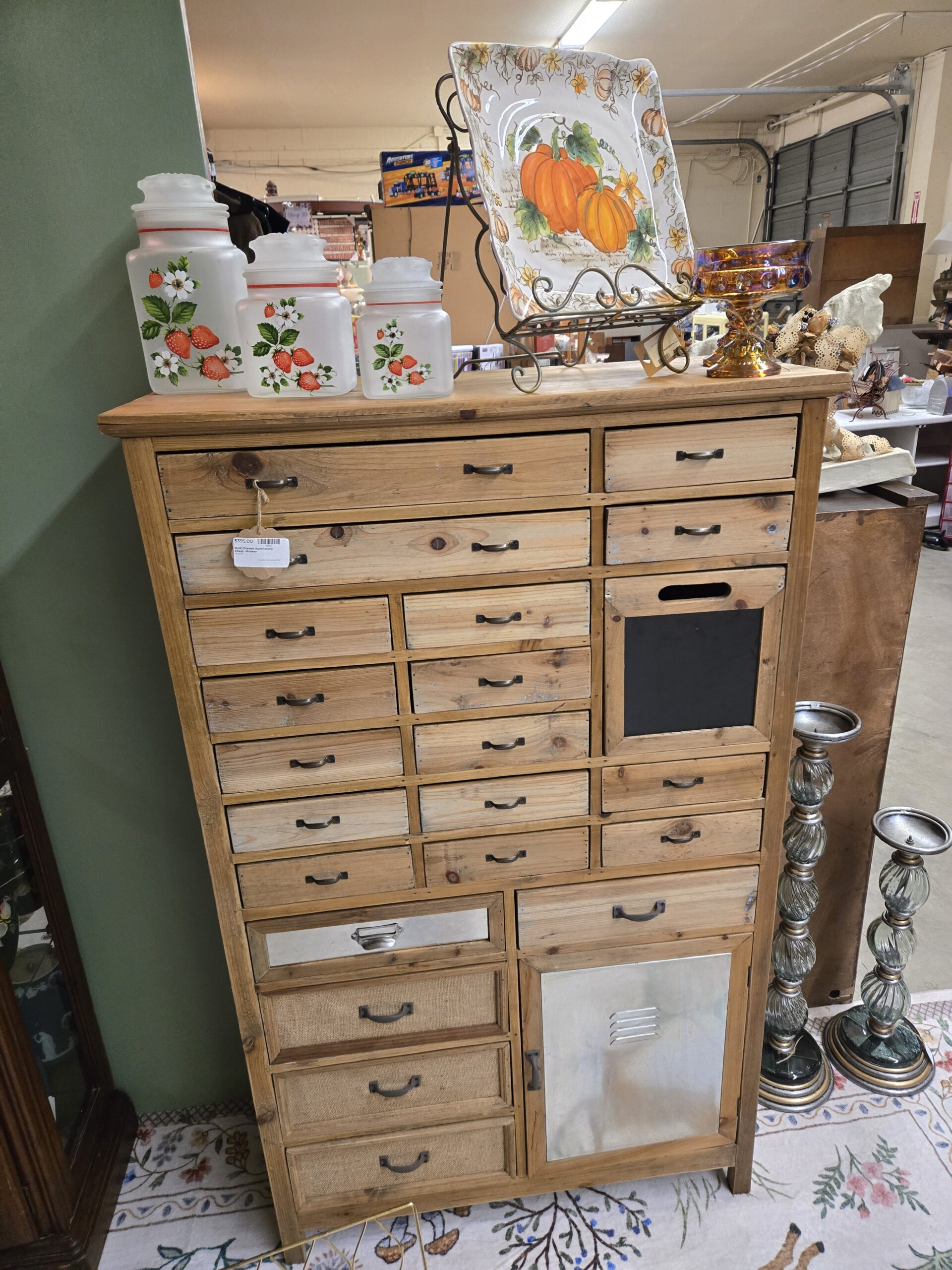 Multi-drawer wood apothecary chest with iron pulls, a chalkboard panel, burlap-front drawers, a metal-front drawer with a bin pull, and a galvanized metal door with vents, topped by three frosted glass canisters painted with strawberries, a pumpkin serving platter on a scrolled metal easel, and an amber carnival glass footed compote, beside two tall glass and silver pillar candle stands at Shop 55, Pacific Antiques Mall in Tacoma, WA.