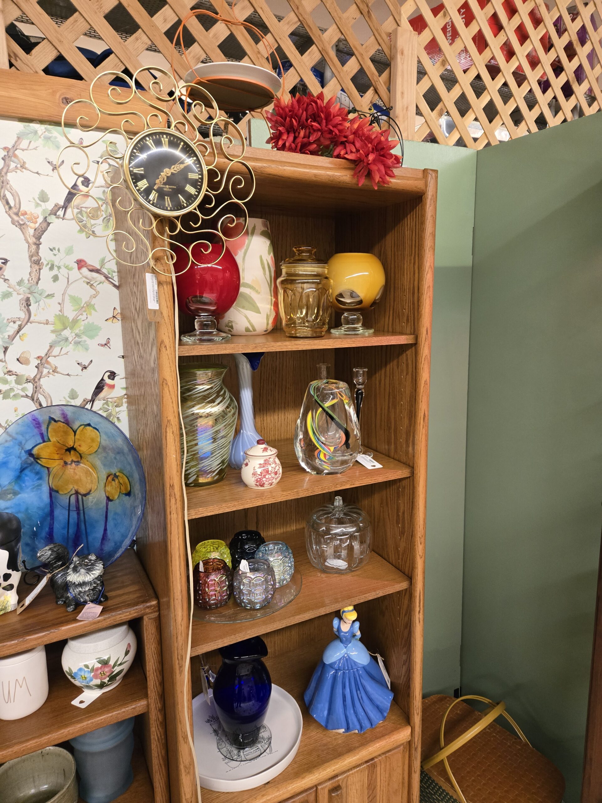 Gold scrollwork wall clock with a black face and Roman numerals, red and yellow cased glass bowls on clear pedestal feet, a painted art pottery vase with red flowers, an amber glass canister, an iridescent swirl art glass vase, a pale blue stretched glass bud vase, a small red floral transferware covered jar, a clear sculptural vase with rainbow ribbons, a black and clear glass candlestick, iridescent votive holders in amethyst, cobalt, yellow-green, and pale blue on a glass tray, a clear glass pumpkin jar, a cobalt glass vase, and a Cinderella figurine in a blue gown at Shop 55, Pacific Antiques Mall in Tacoma, WA.