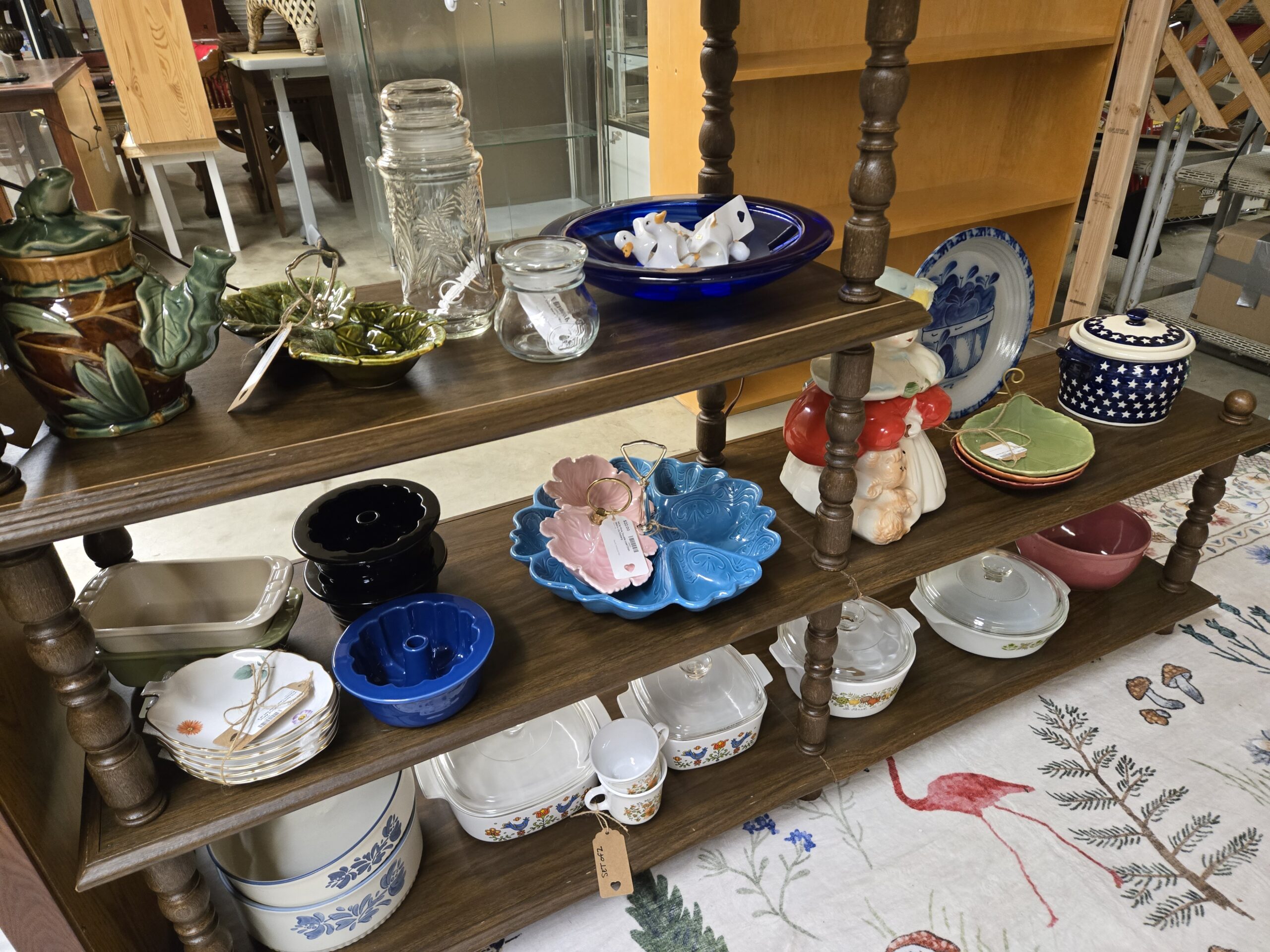 Leaf-wrapped teapot with a frog on the lid in majolica-style green and brown glazes, green leaf serving dishes with a center handle, a clear pressed glass canister with a wheat pattern, a cobalt blue glass bowl holding white ceramic ducks, a blue and white stoneware plate with an apple basket, a Polish pottery covered pot with blue stars, a figural cookie jar of a woman in a red cloak with a puppy, green leaf plates, pink and turquoise leaf-shaped serving dishes with brass handles, black and cobalt fluted tube pans, gold-trimmed floral dessert bowls, a pink mixing bowl, Corning Ware Spice of Life and Country Festival casseroles and mugs, and Pfaltzgraff Yorktowne bowls at Shop 55, Pacific Antiques Mall in Tacoma, WA.