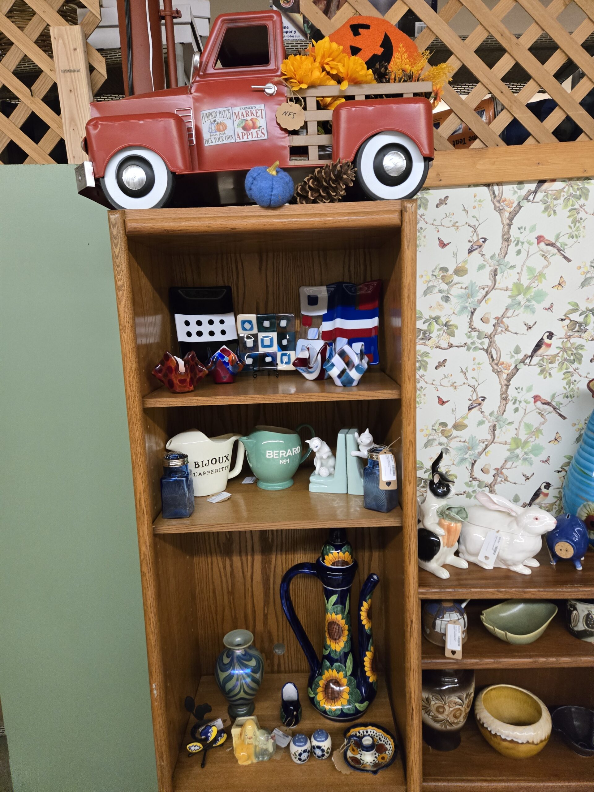 Fused glass plates in black with white dots, teal and blue checkerboard, and red, white, and blue stripes, fused glass votive holders in red, cobalt, and checkered blue, a cream Bijoux L'Apperitif pitcher, a mint green Berard No1 pitcher, white cat bookends on mint bases, cobalt blue glass shakers with metal lids, an iridescent pulled-feather art glass vase, a cobalt ewer painted with sunflowers, a yellow puppy planter, blue and white salt and pepper shakers, a matching painted chamberstick, and a butterfly ornament, beneath a red metal farm truck display with autumn flowers at Shop 55, Pacific Antiques Mall in Tacoma, WA.