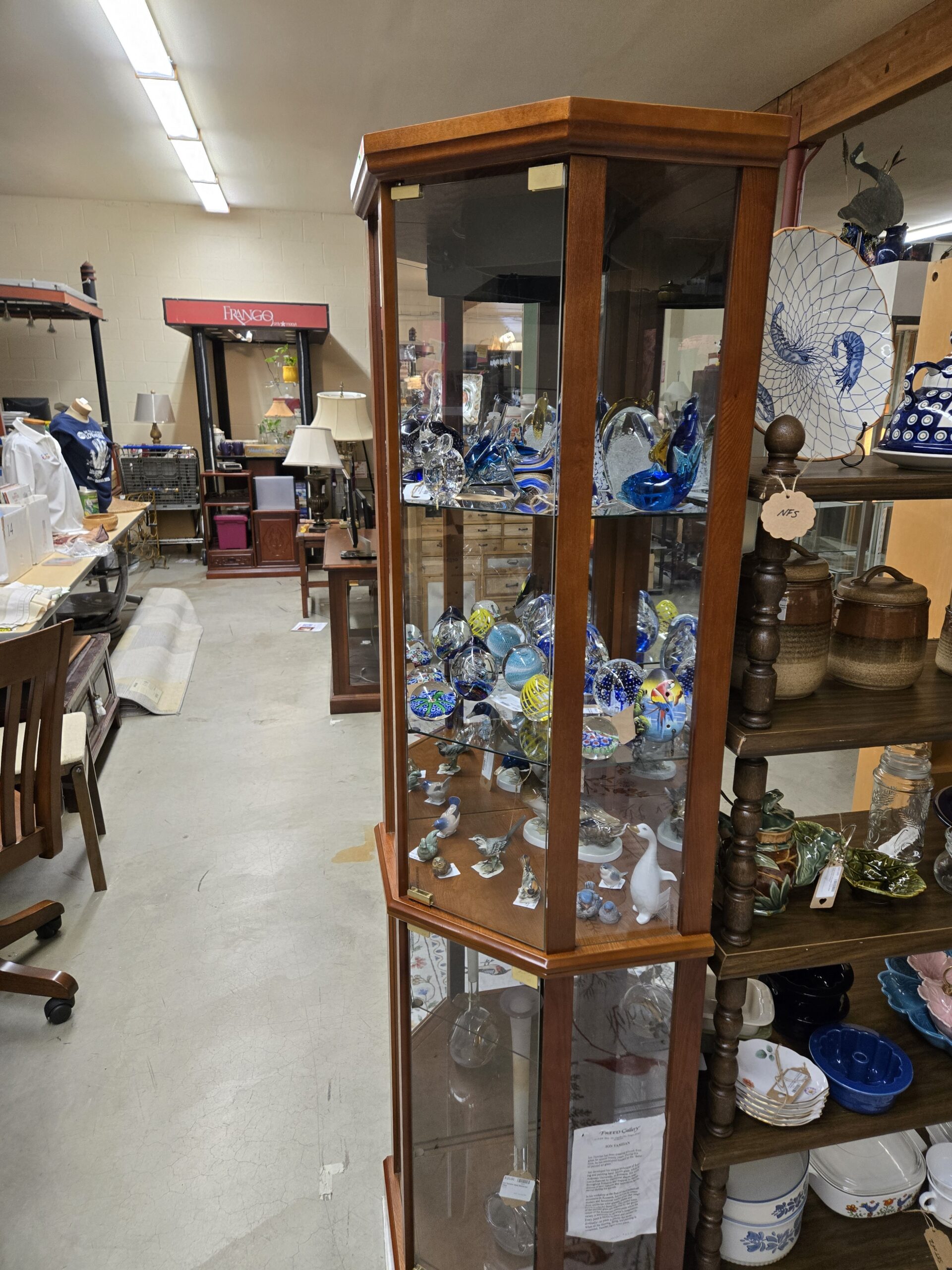 Hexagonal wood and glass curio cabinet holding blue art glass fish and dolphin sculptures, a clear glass bird, millefiori paperweights, controlled-bubble paperweights, yellow ribbon swirl paperweights, an egg-shaped paperweight painted with a parrot, porcelain songbird and bluebird figurines, a small owl figurine, a white goose figurine, and a tall clear glass bud vase at Shop 55, Pacific Antiques Mall in Tacoma, WA.